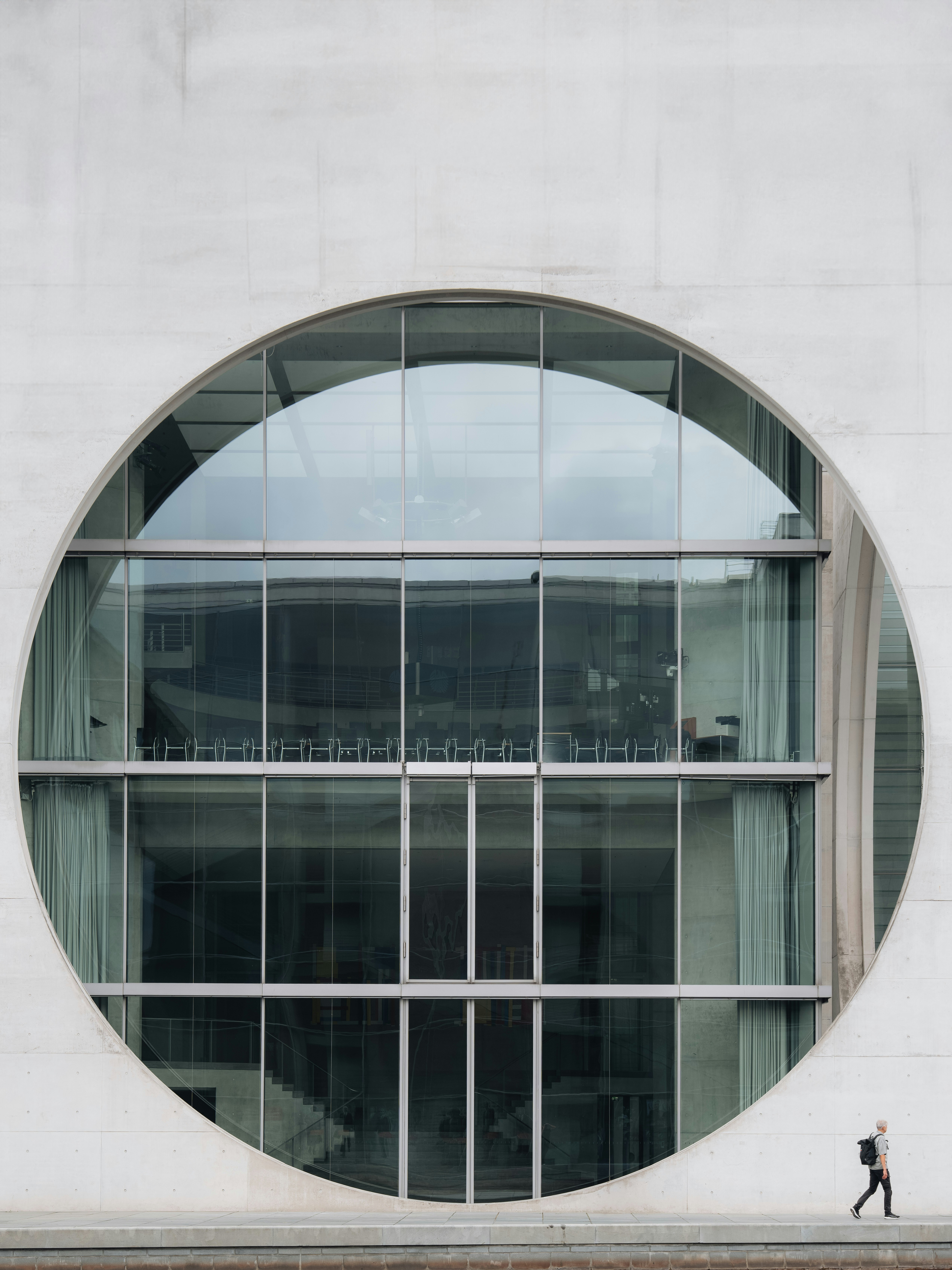 A person walking past a large circular glass window on a concrete building
