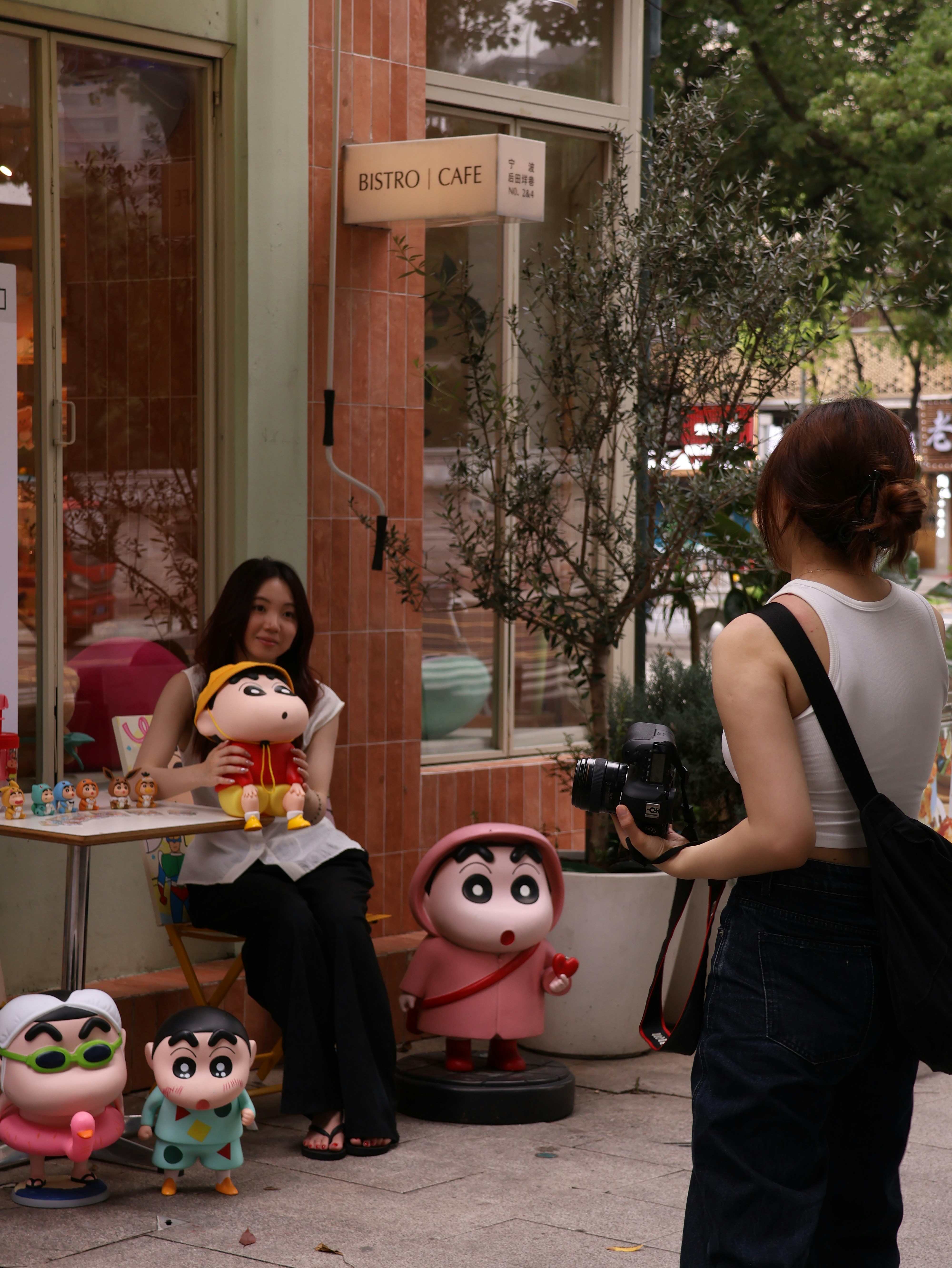 A woman photographing another woman holding a cartoon character doll outside a cafe