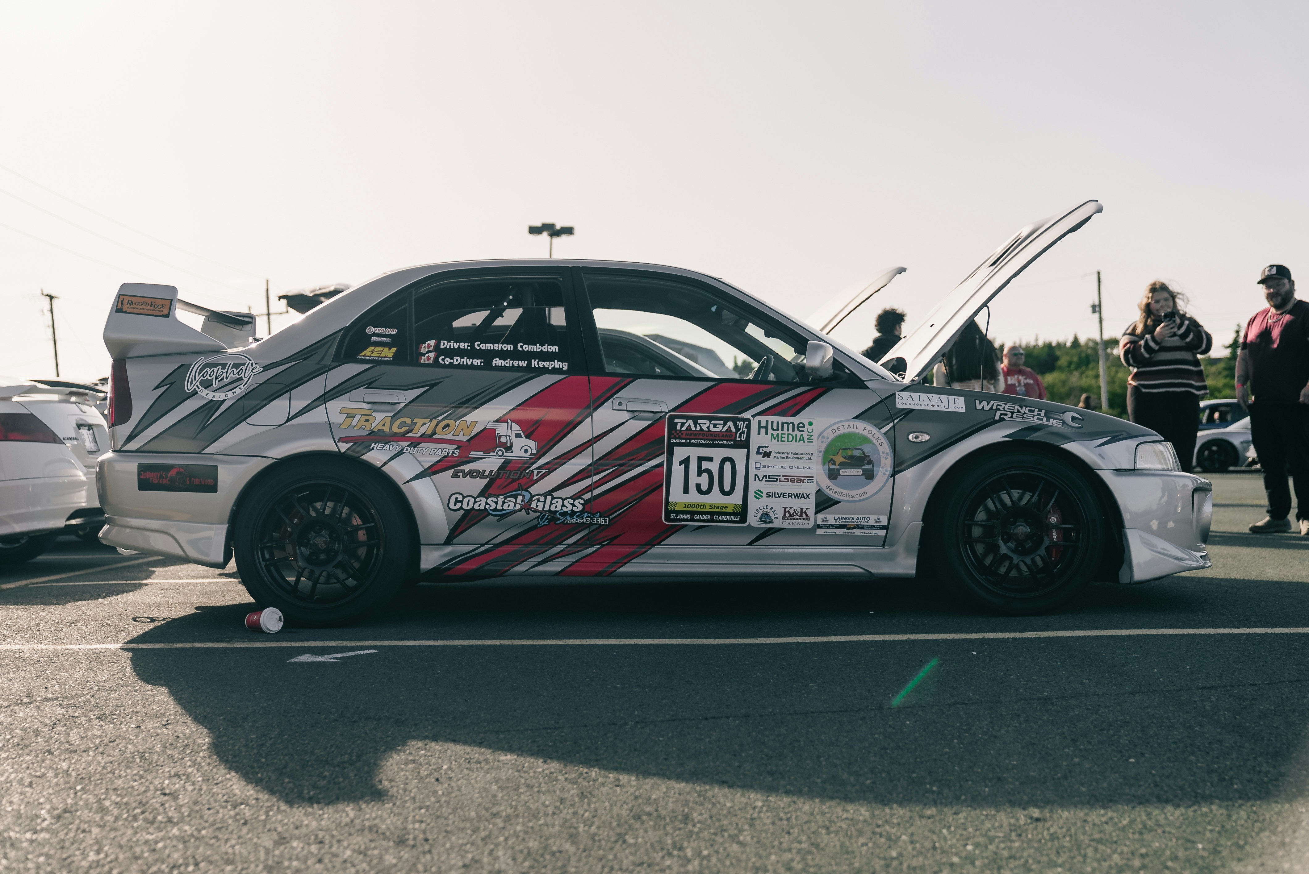 A silver rally car with racing decals and an open hood in a parking lot