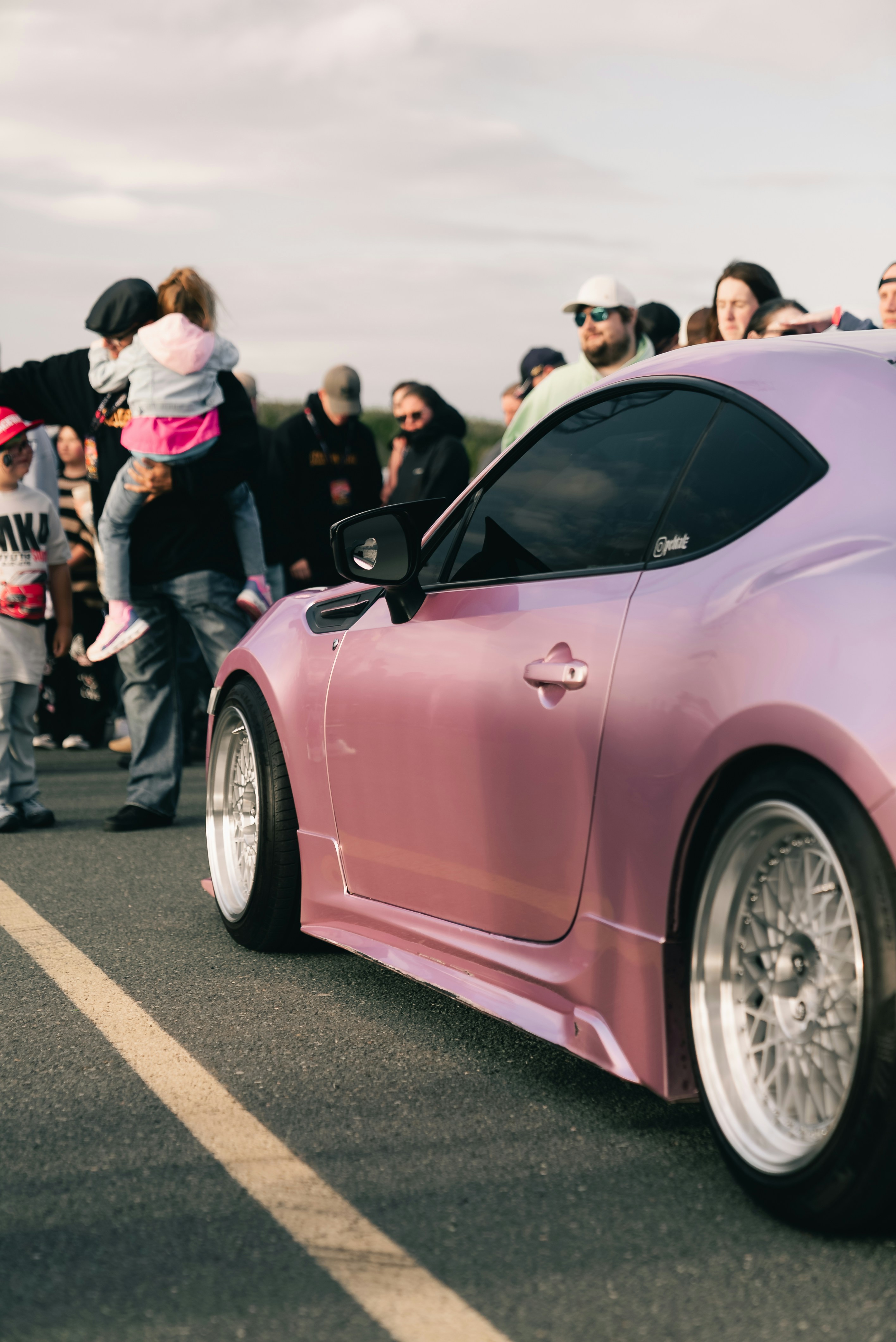 A pink sports car parked on asphalt with a crowd of people nearby