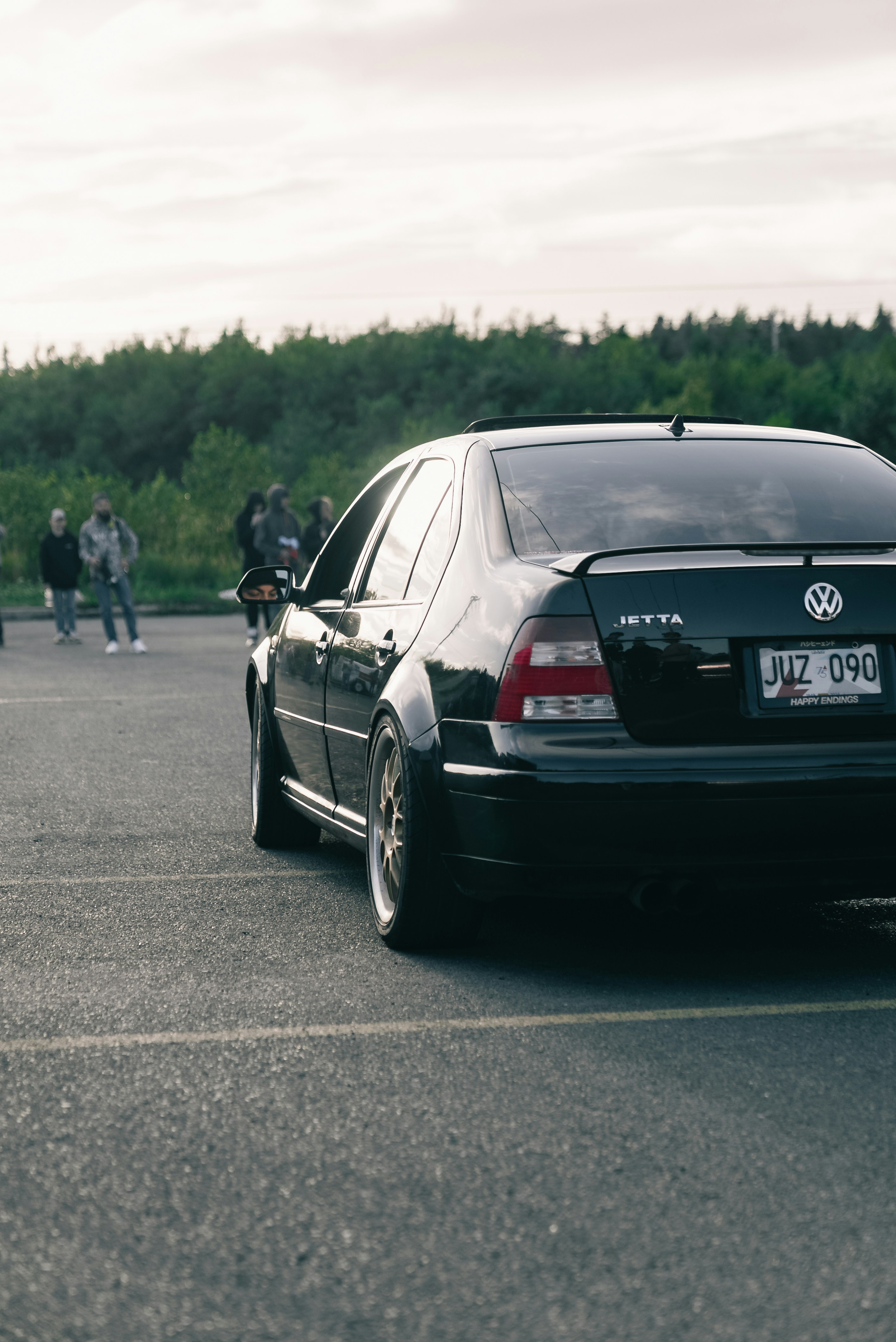 A black sedan parked in a lot with people standing in the background