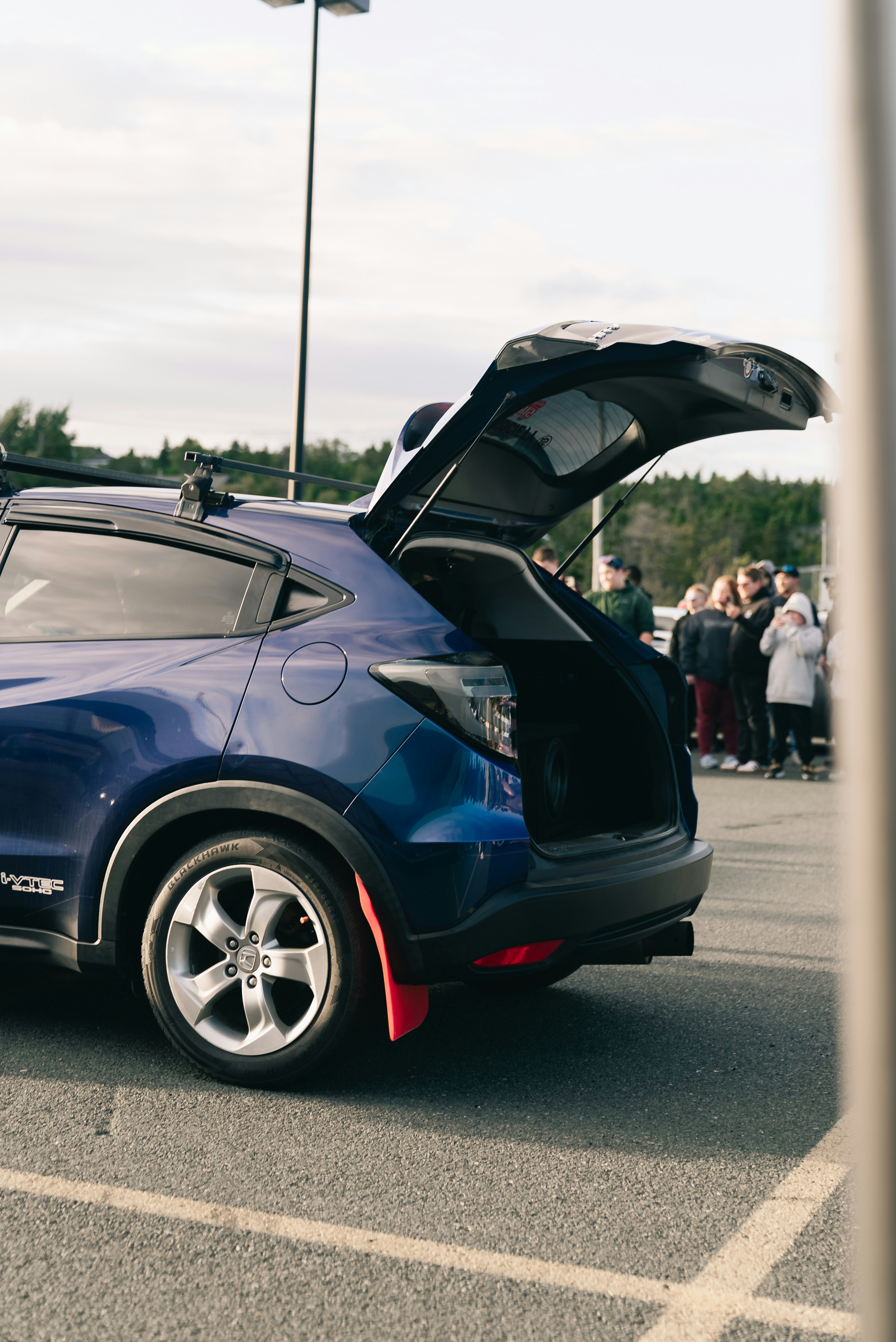 A blue hatchback with an open trunk parked in a lot with people