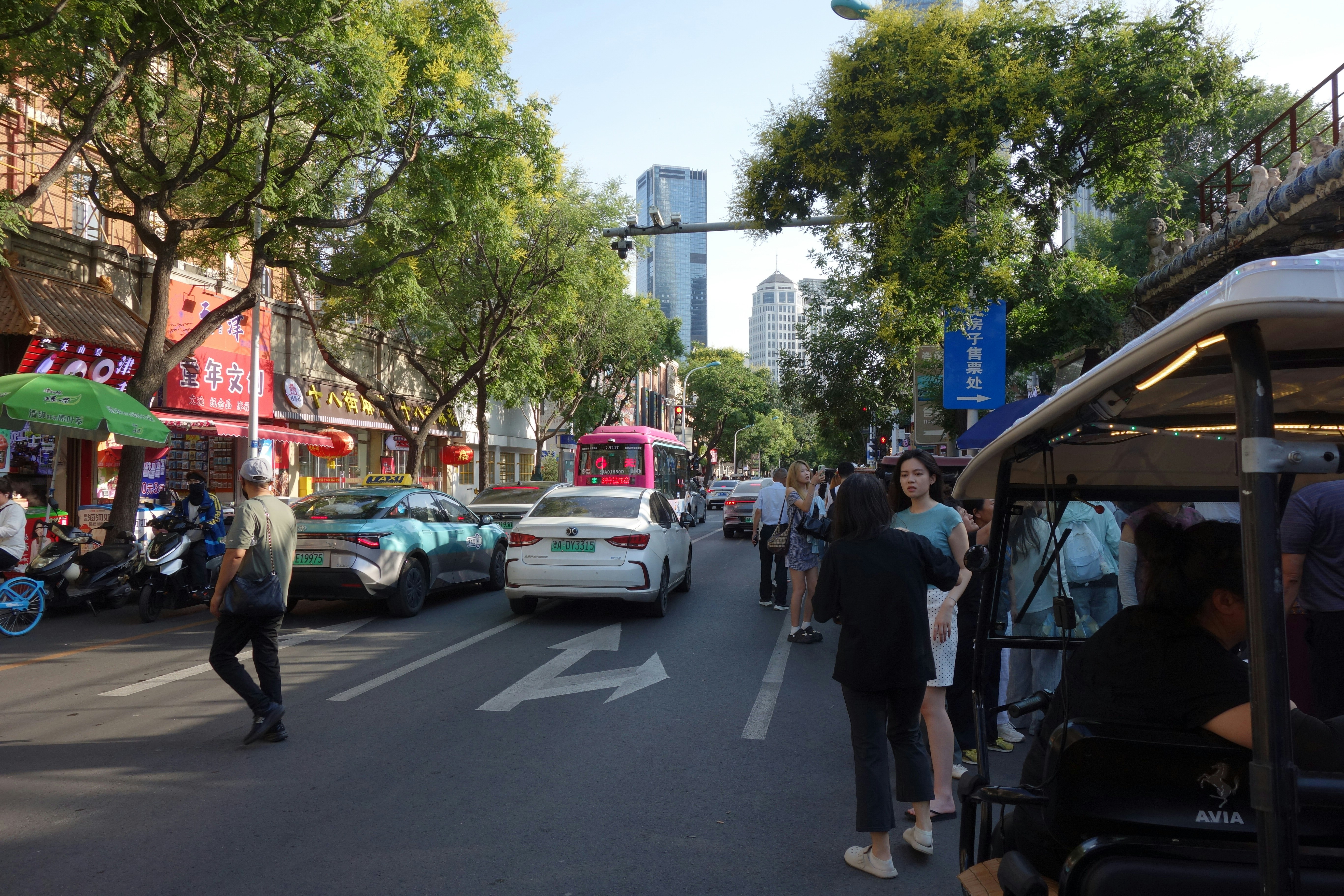 Pessoas caminhando e paradas em uma rua movimentada da cidade, com carros e árvores