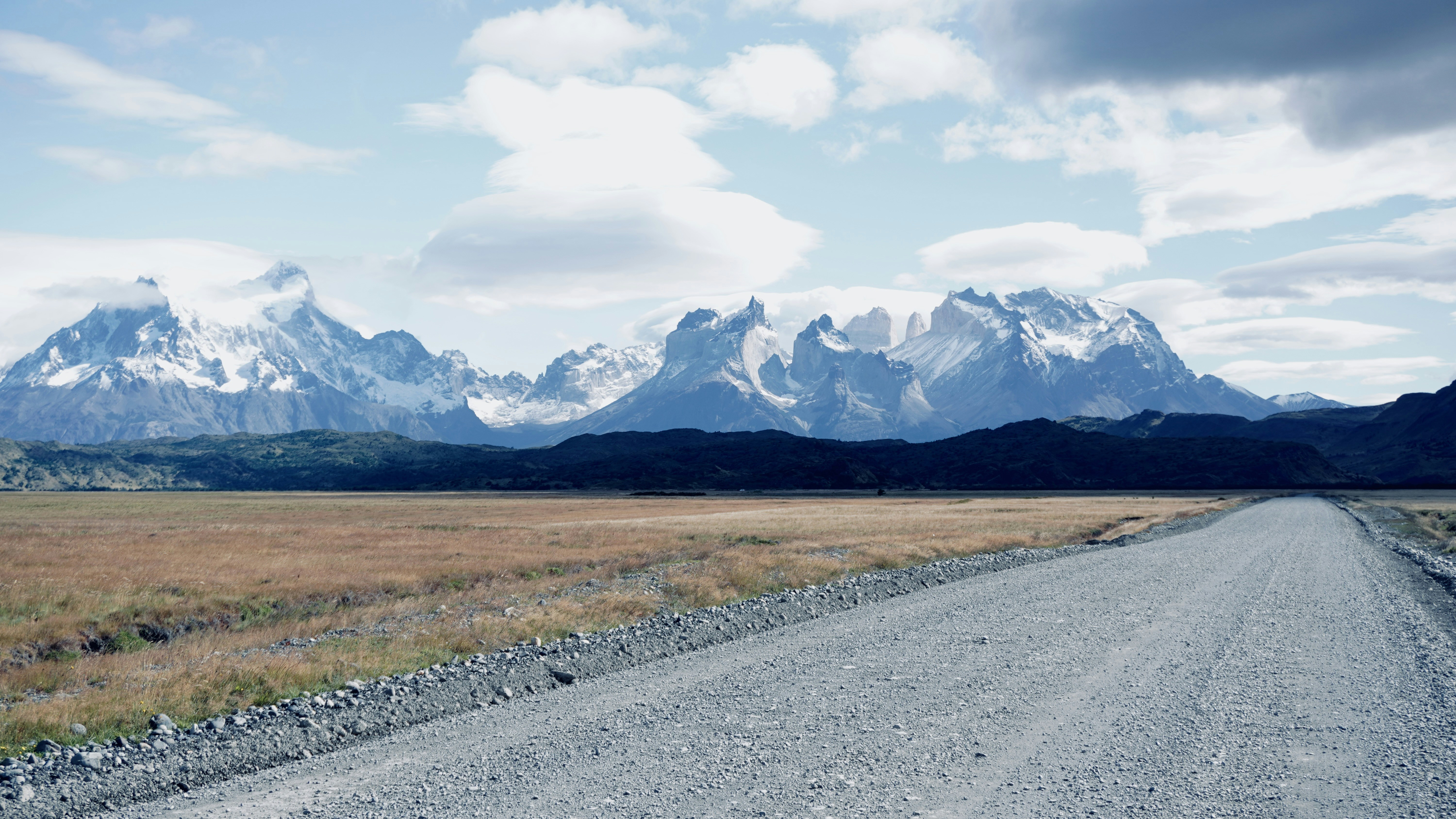 A gravel road leading toward snow-capped mountains under a blue sky with clouds