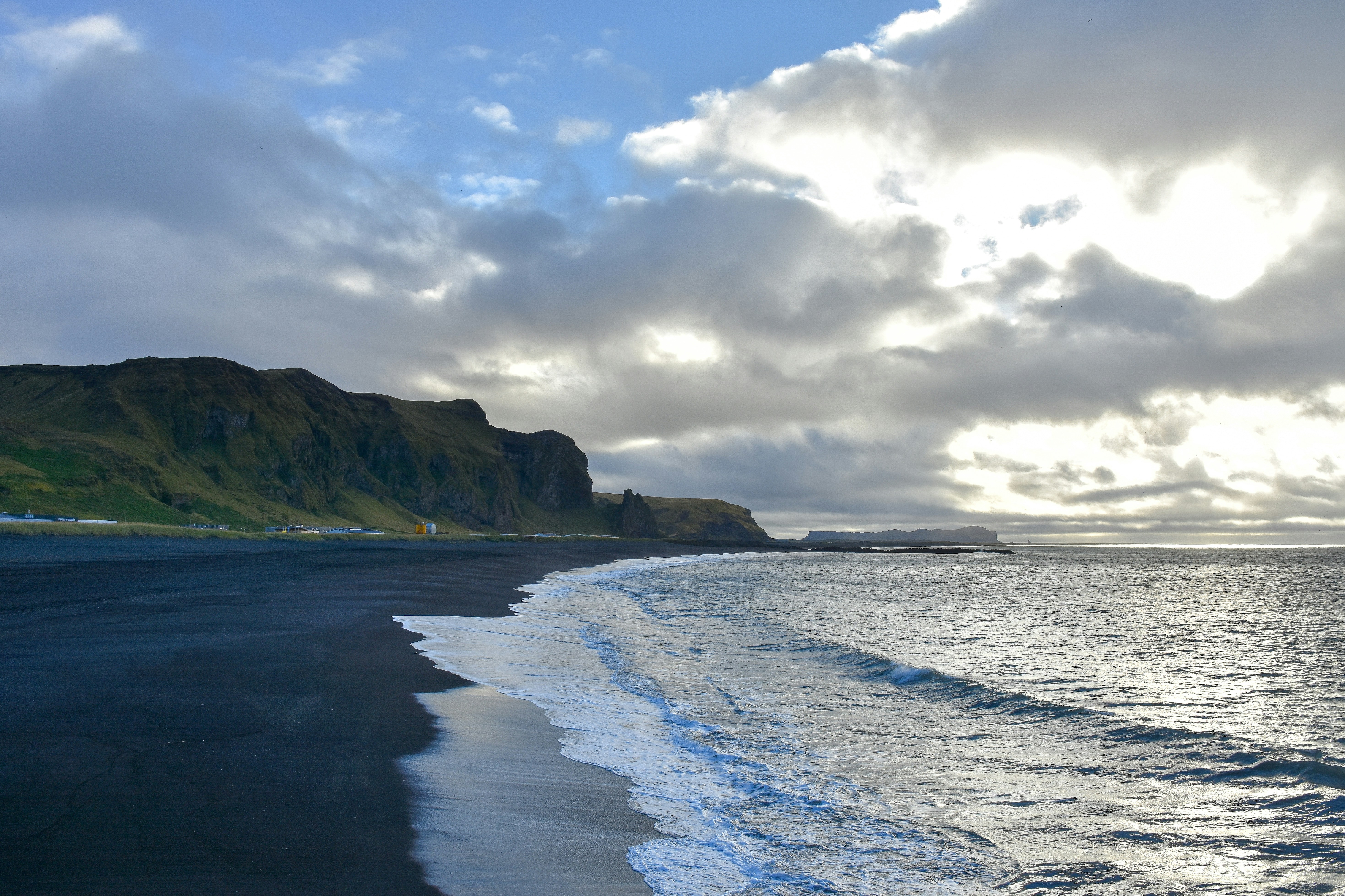 Waves washing onto a black sand beach below green cliffs under a cloudy sky