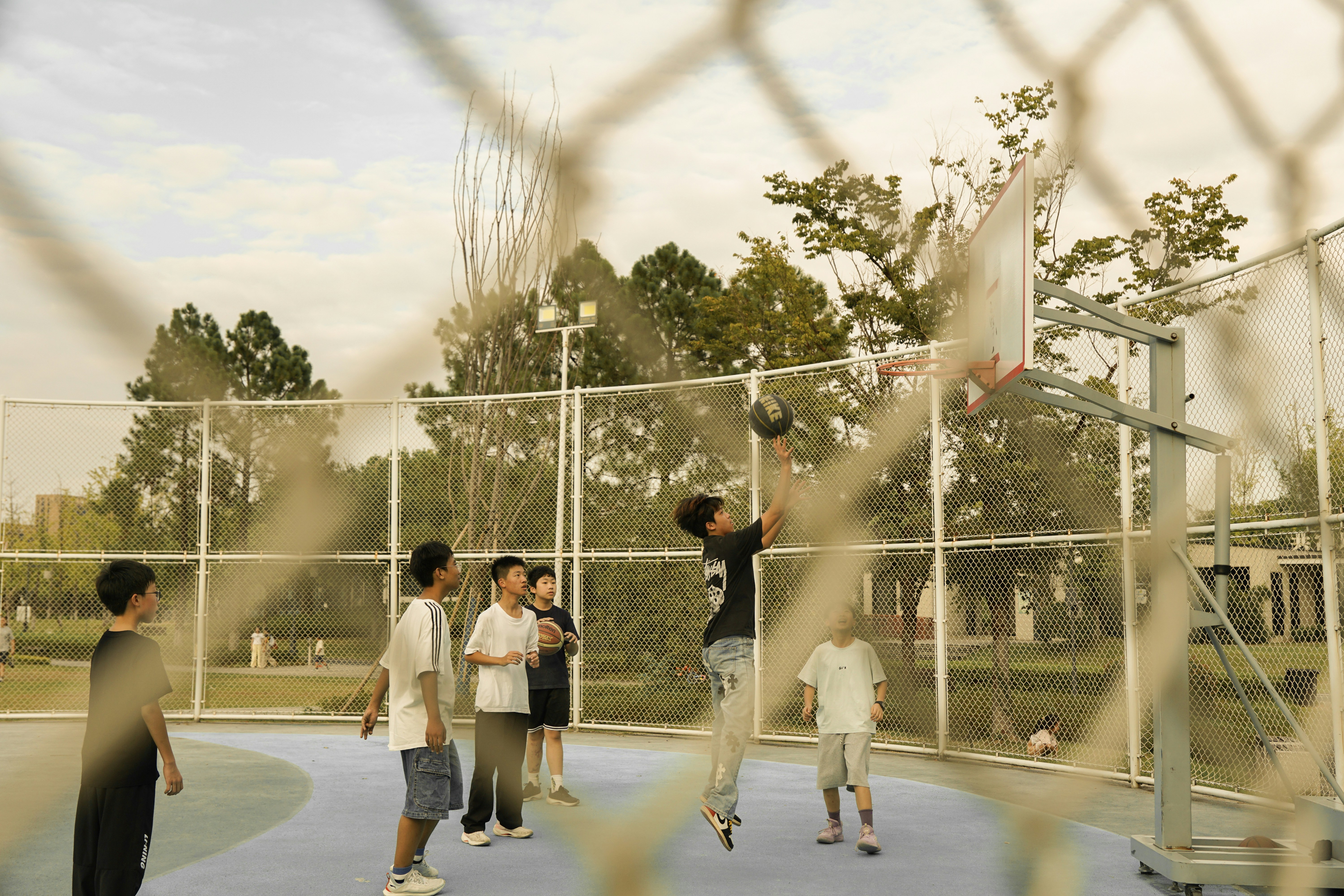 A group of teenagers playing basketball on an outdoor court behind a chain-link fence