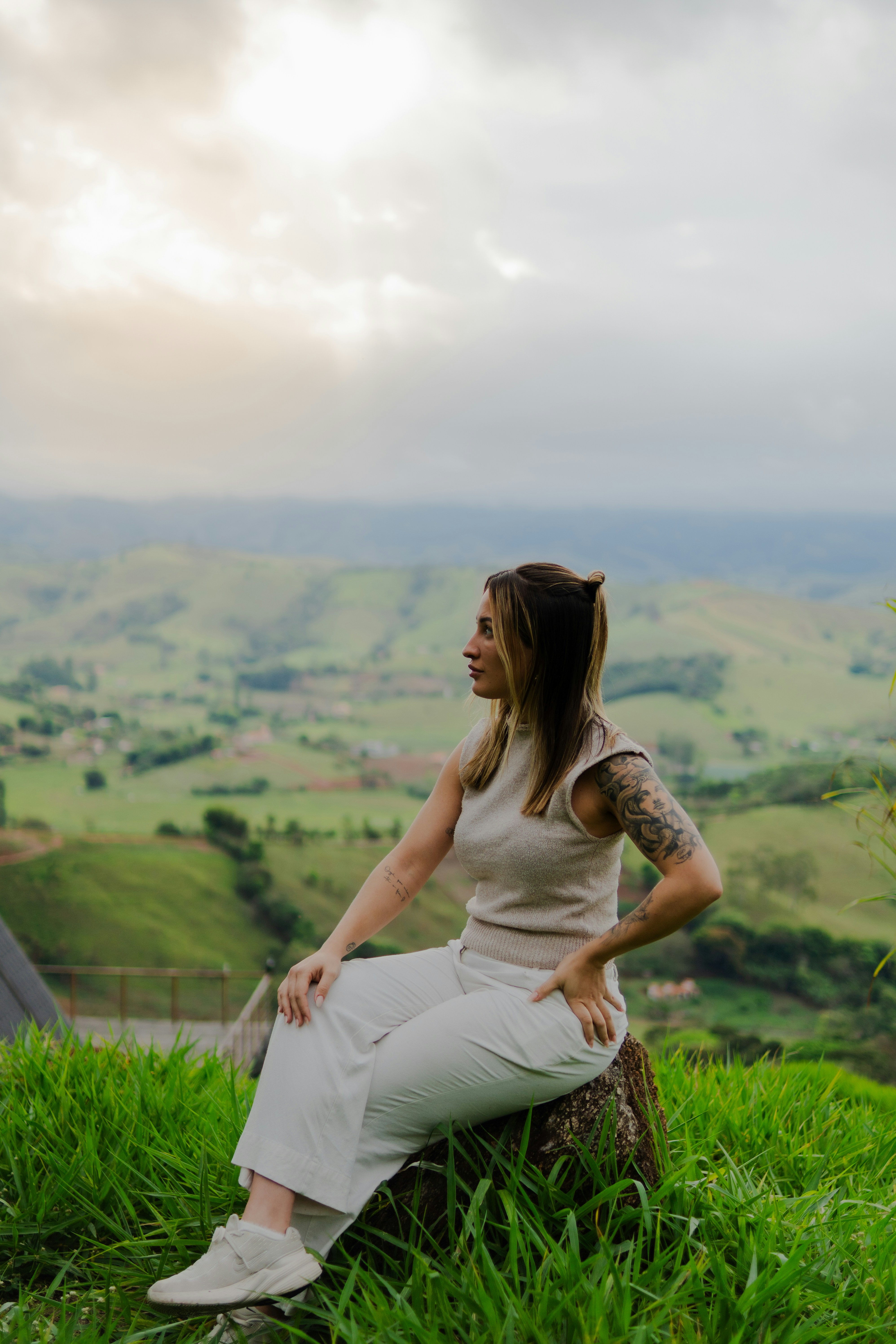 A woman with tattoos sitting on a rock in a grassy field overlooking hills