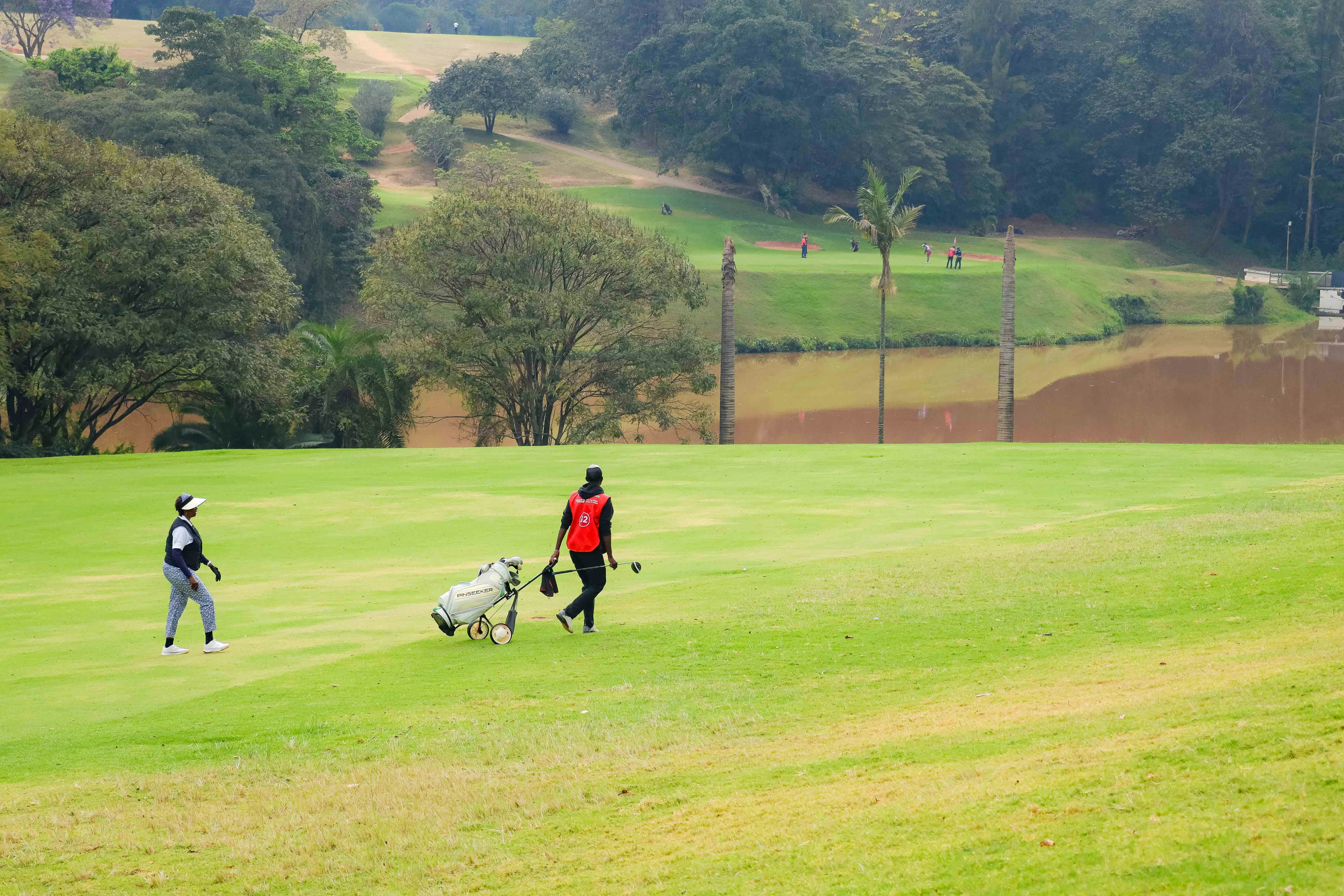 A golfer and a caddie walking across a green golf course near a pond