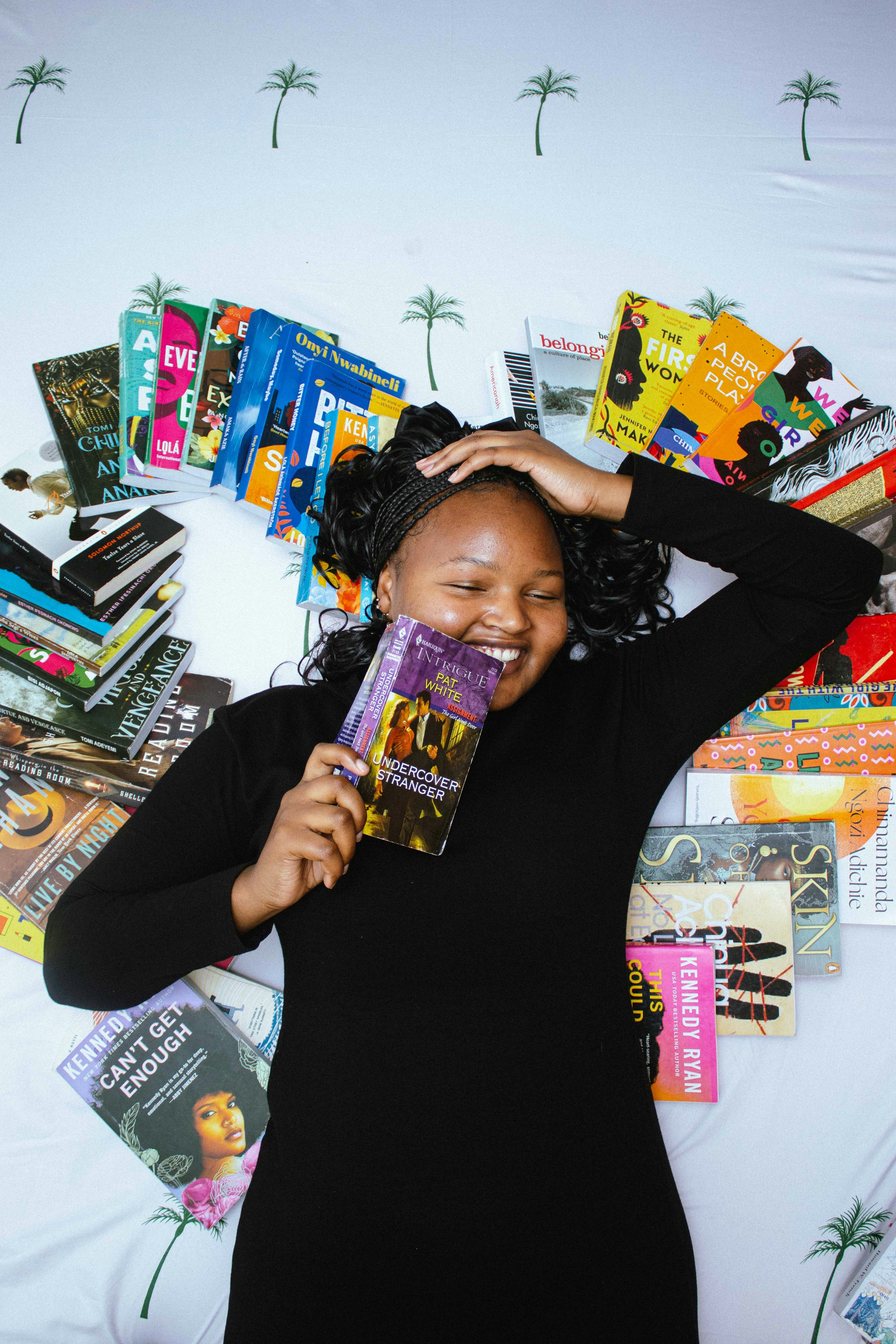 A smiling woman in a black dress lying on a white surface surrounded by books