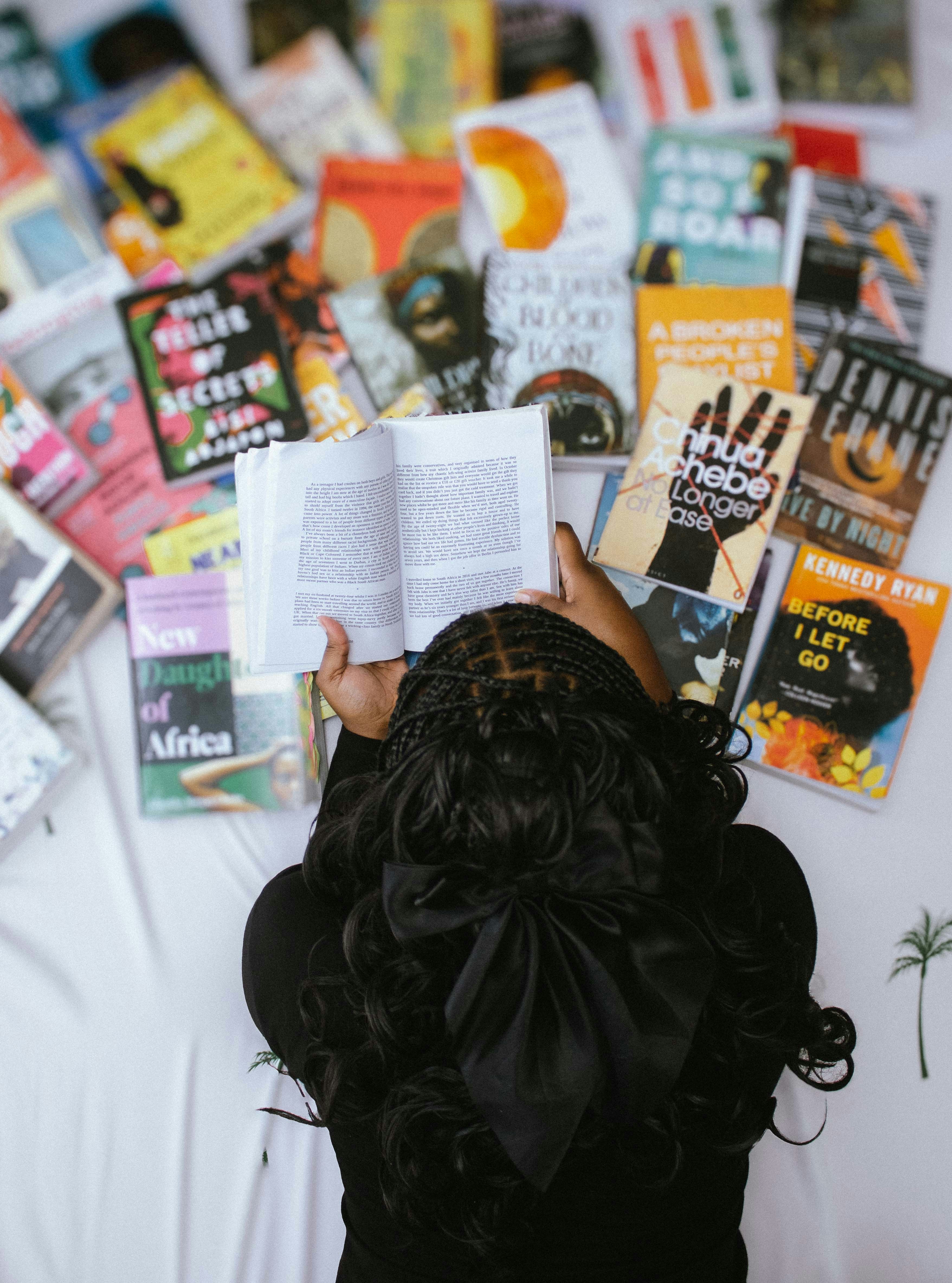 A person with braided hair reading a book surrounded by many other books