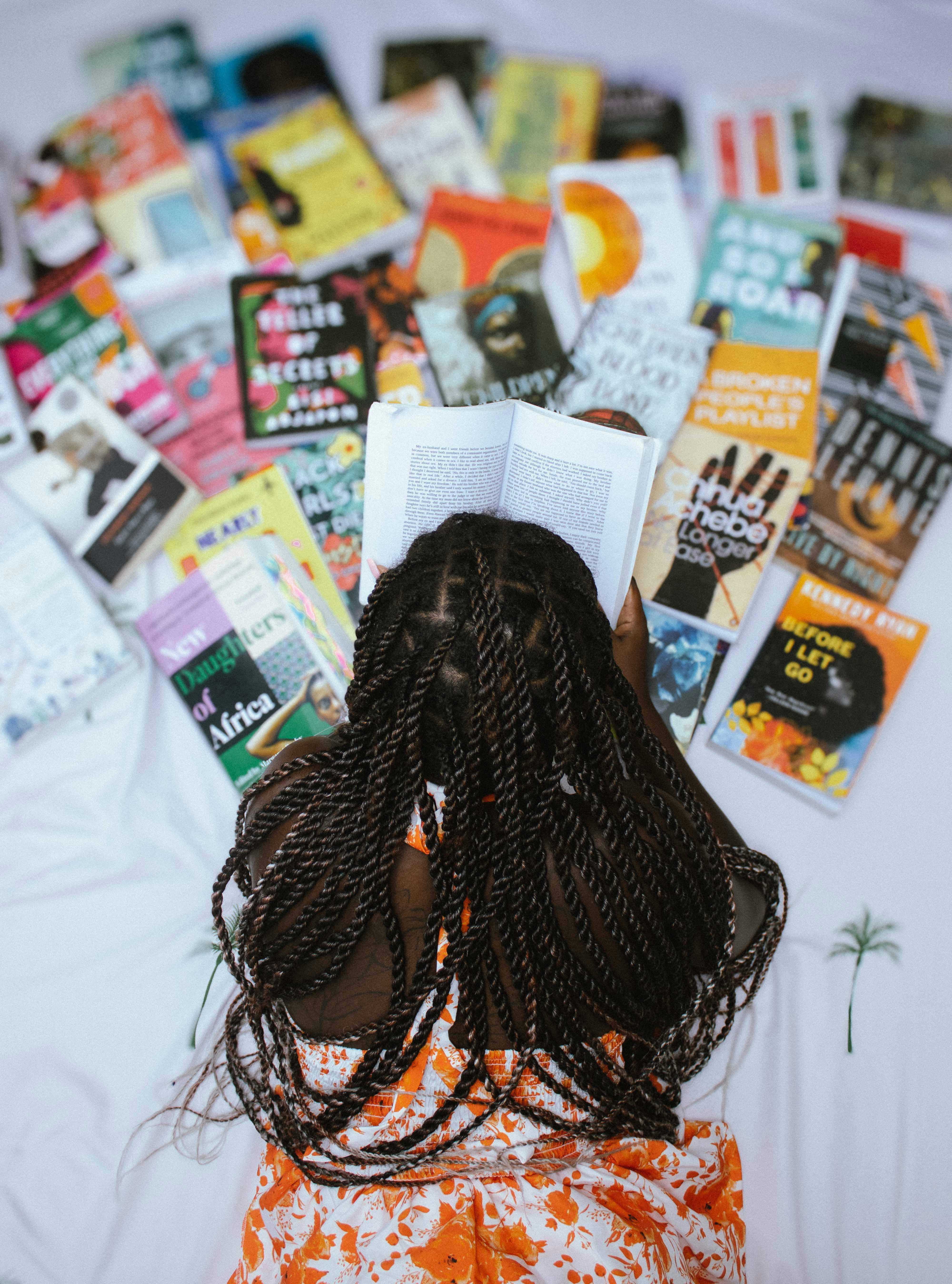 A person with braided hair reading a book surrounded by many other books