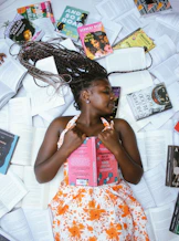 A woman with braided hair lying on a floor covered in many open books