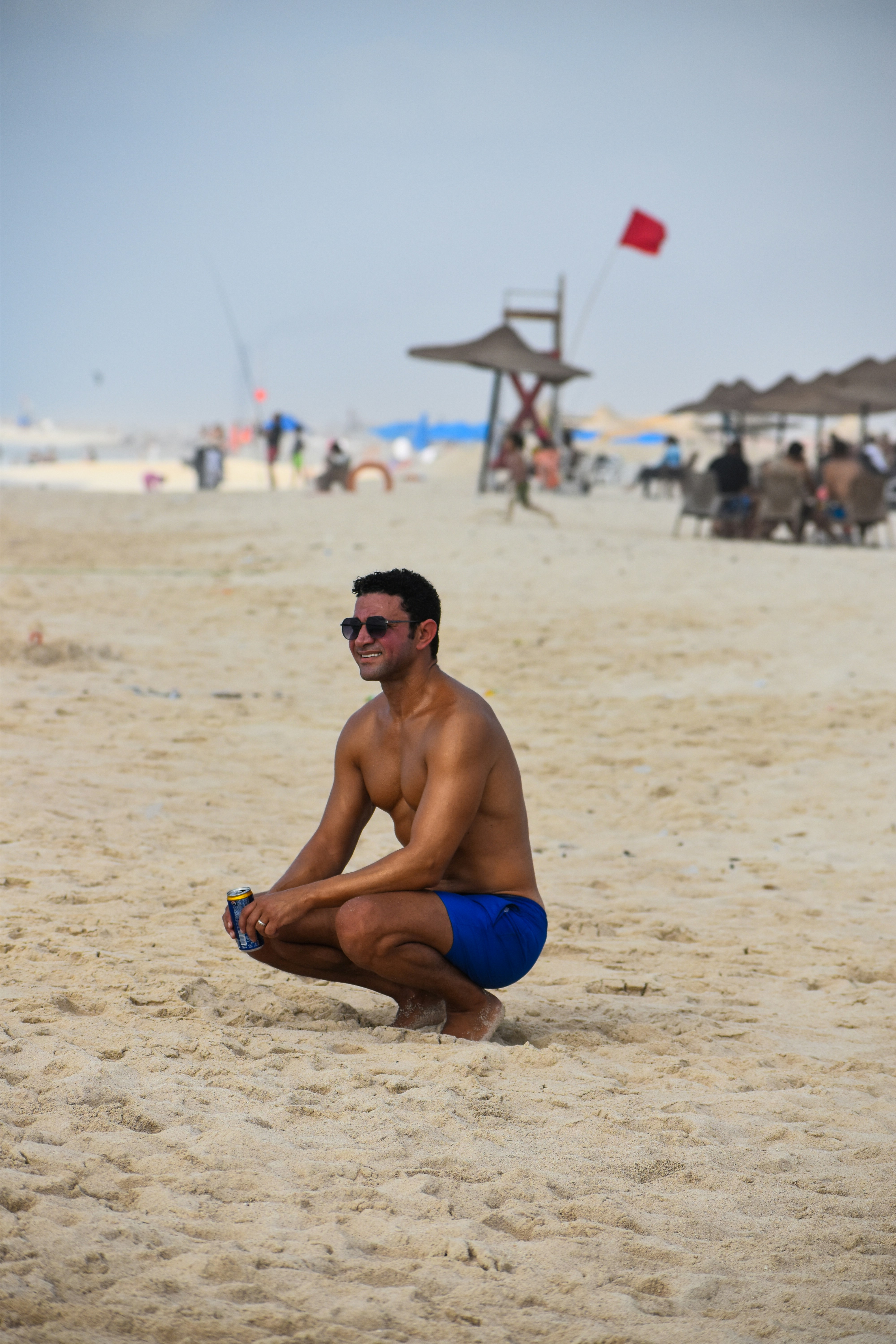 Un homme en maillot de bain bleu et lunettes de soleil accroupi sur une plage de sable