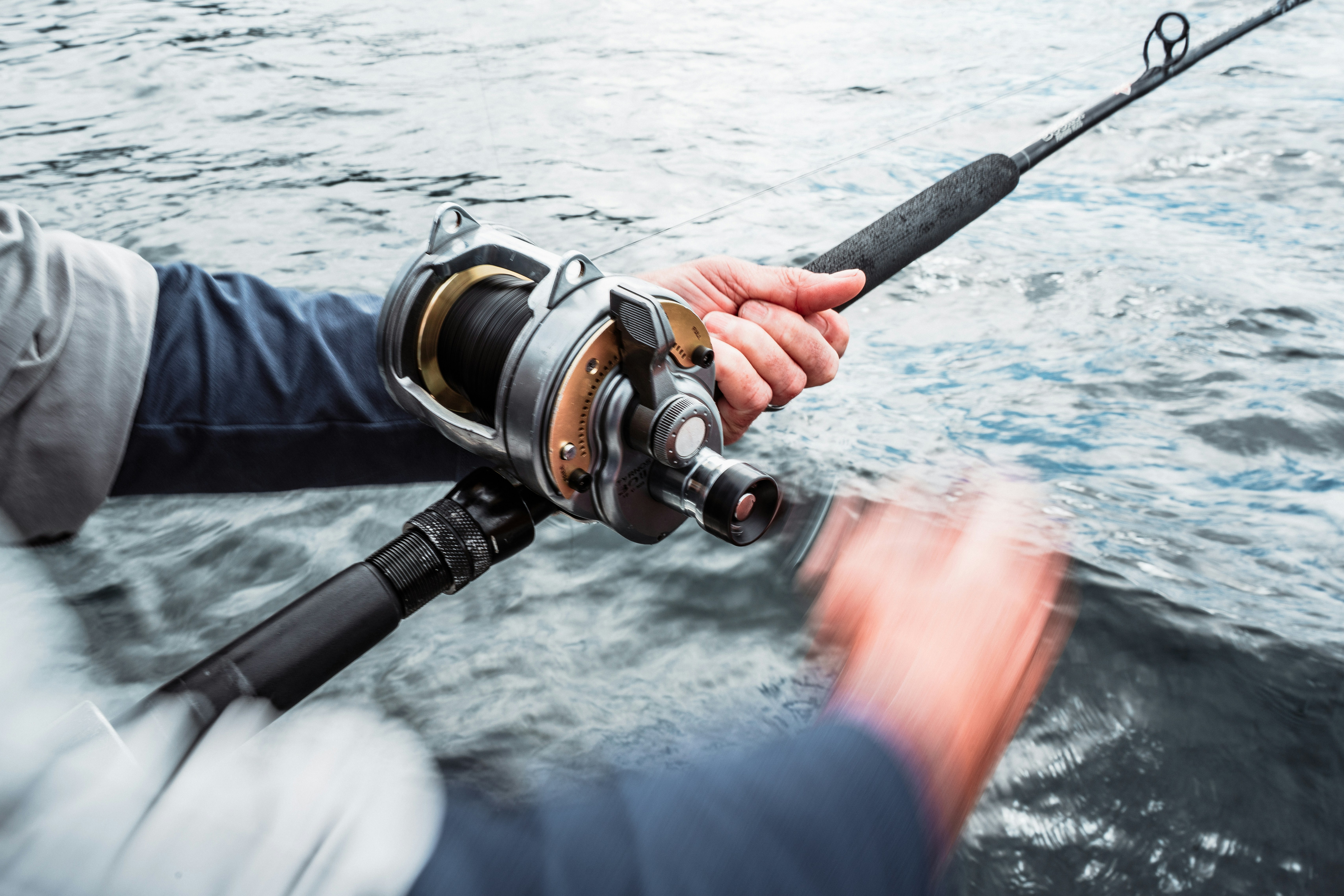 Angler's hands turning the handle of a two-speed conventional reel on a heavy rod bent toward choppy gray water