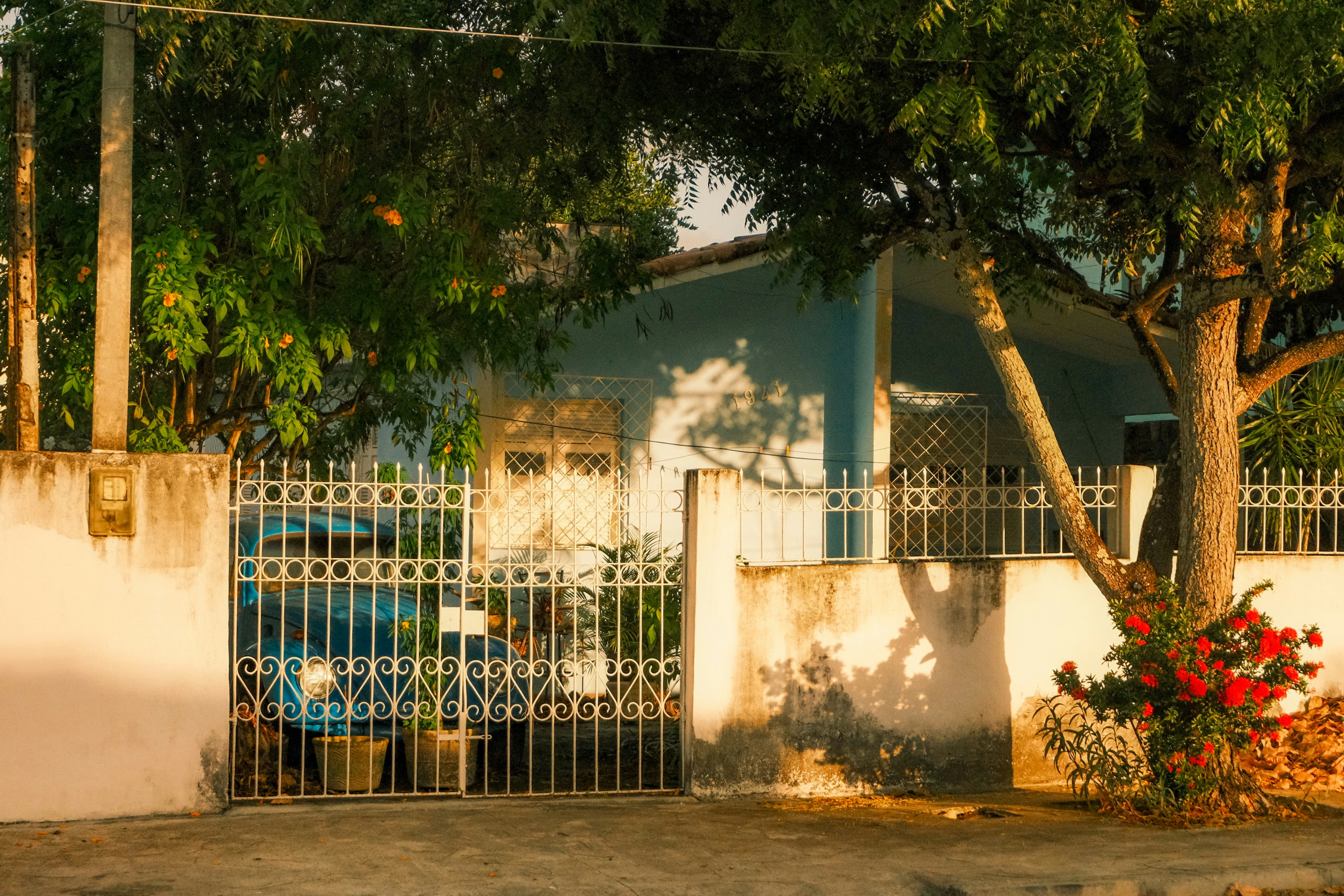 Rumah kecil bercat biru muda di balik tembok dan gerbang besi berukir, disinari matahari sore