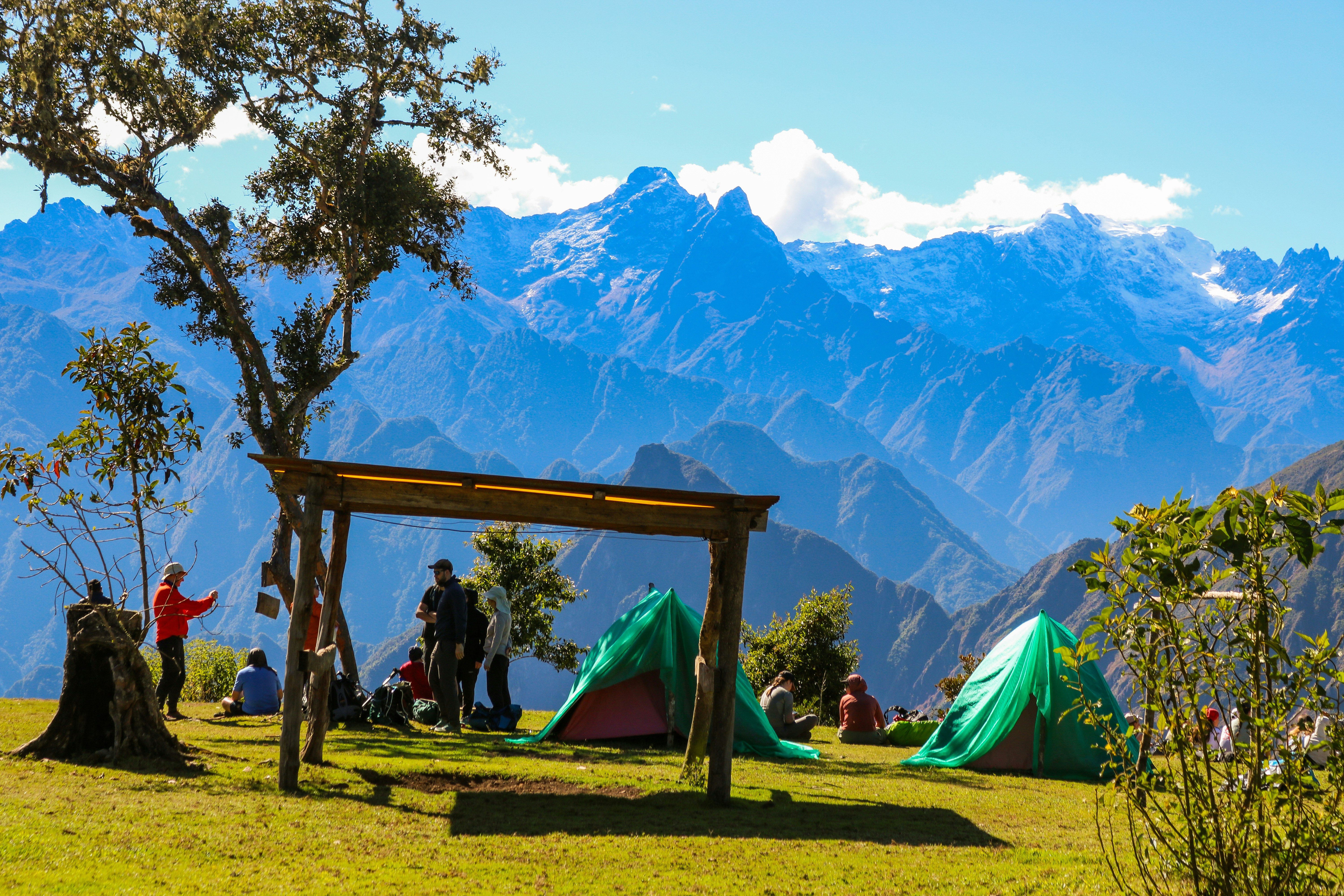 People camping in green tents on a grassy mountain plateau with snow-capped peaks