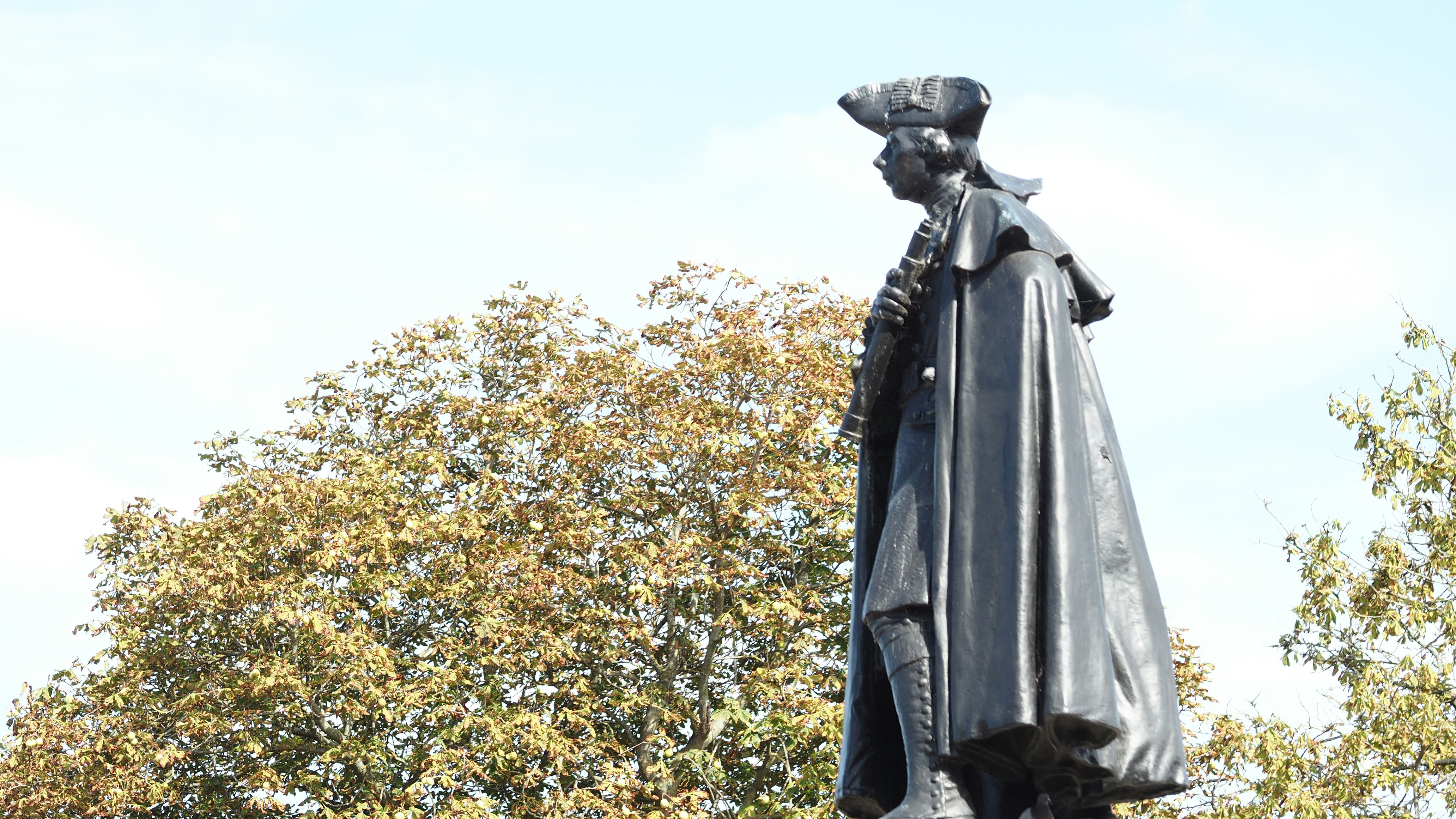 Una statua di bronzo di un uomo con un cappello a tricorno e un mantello davanti agli alberi autunnali