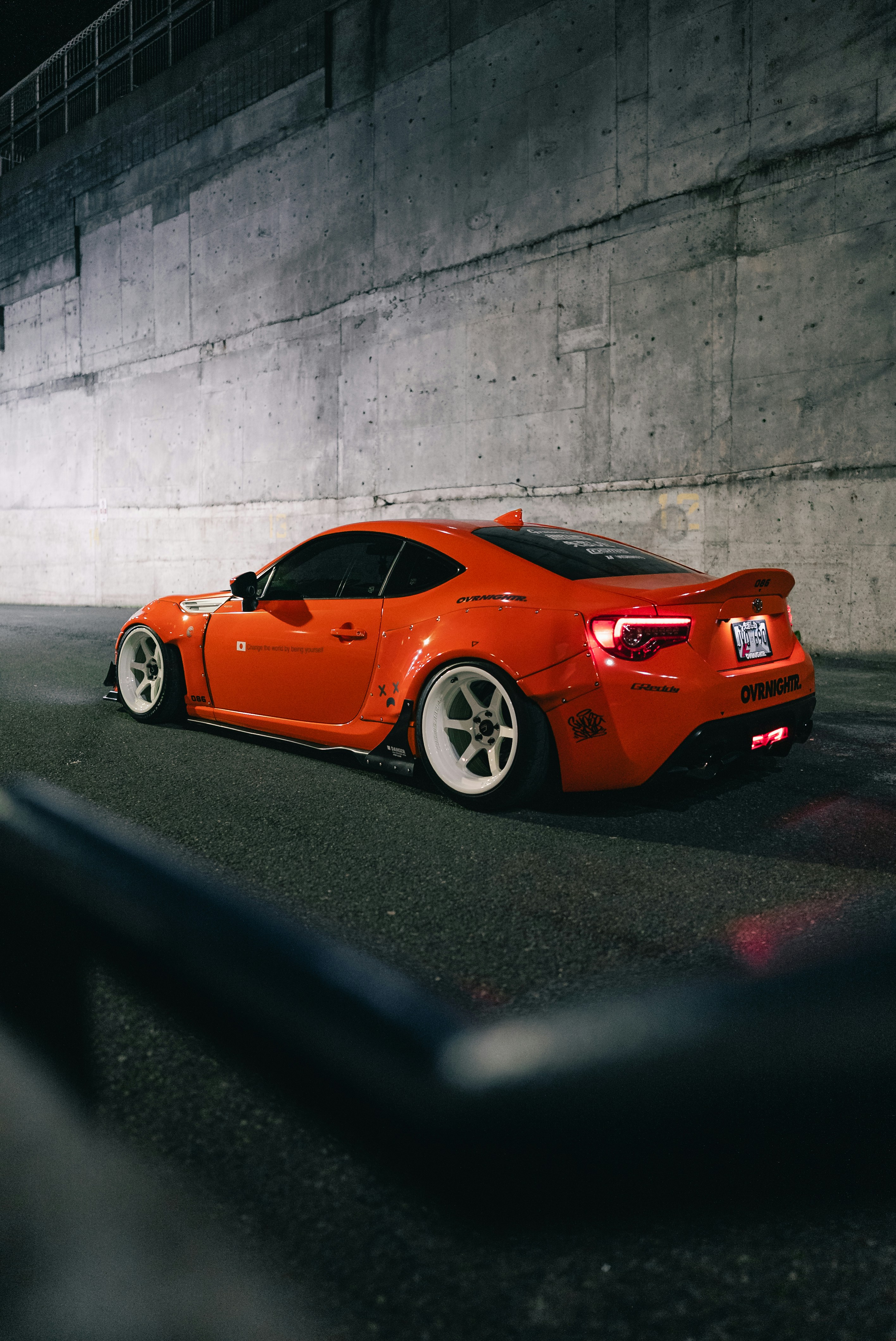 An orange sports car with white wheels parked against a concrete wall at night