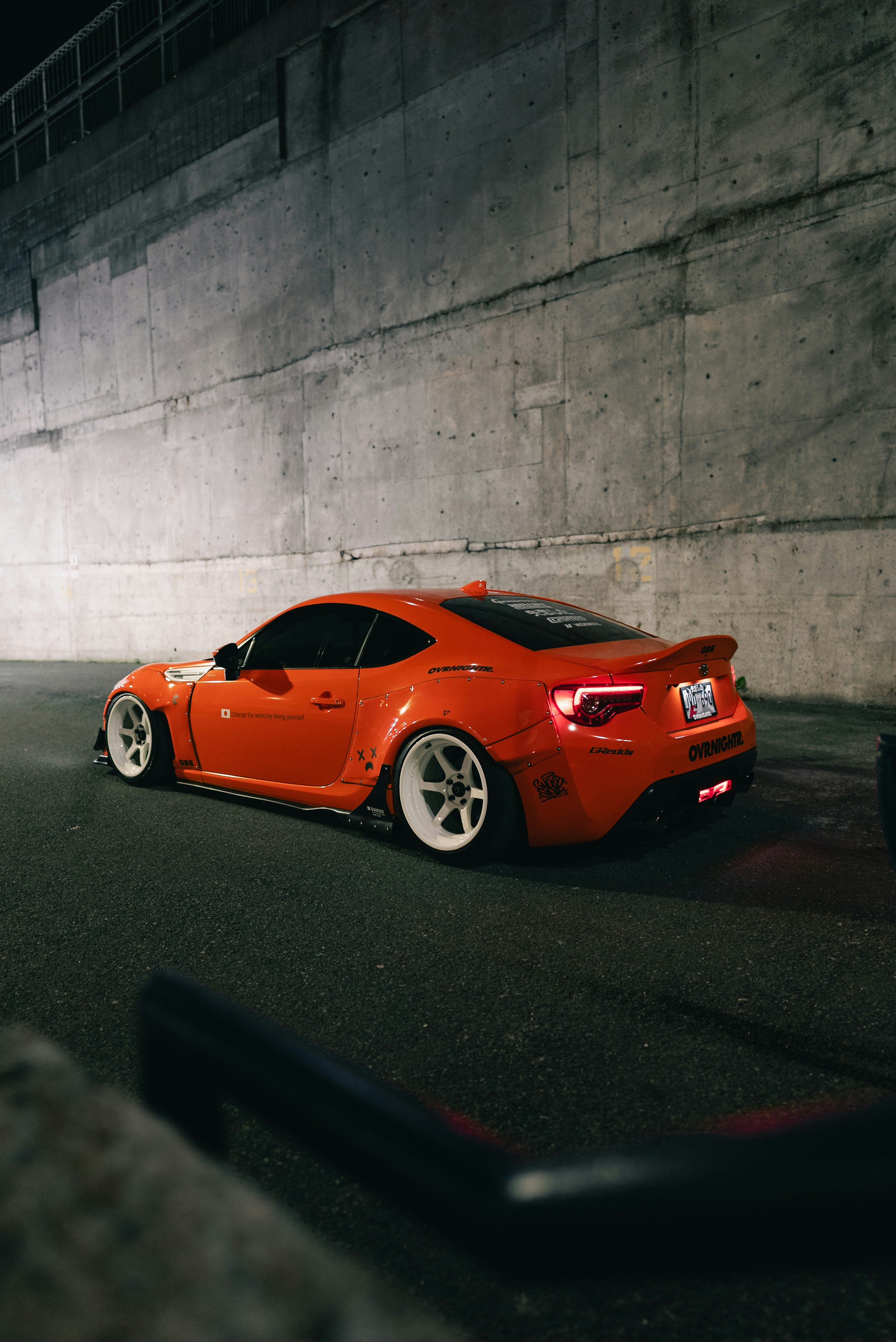 An orange modified sports car with white wheels parked against a concrete wall