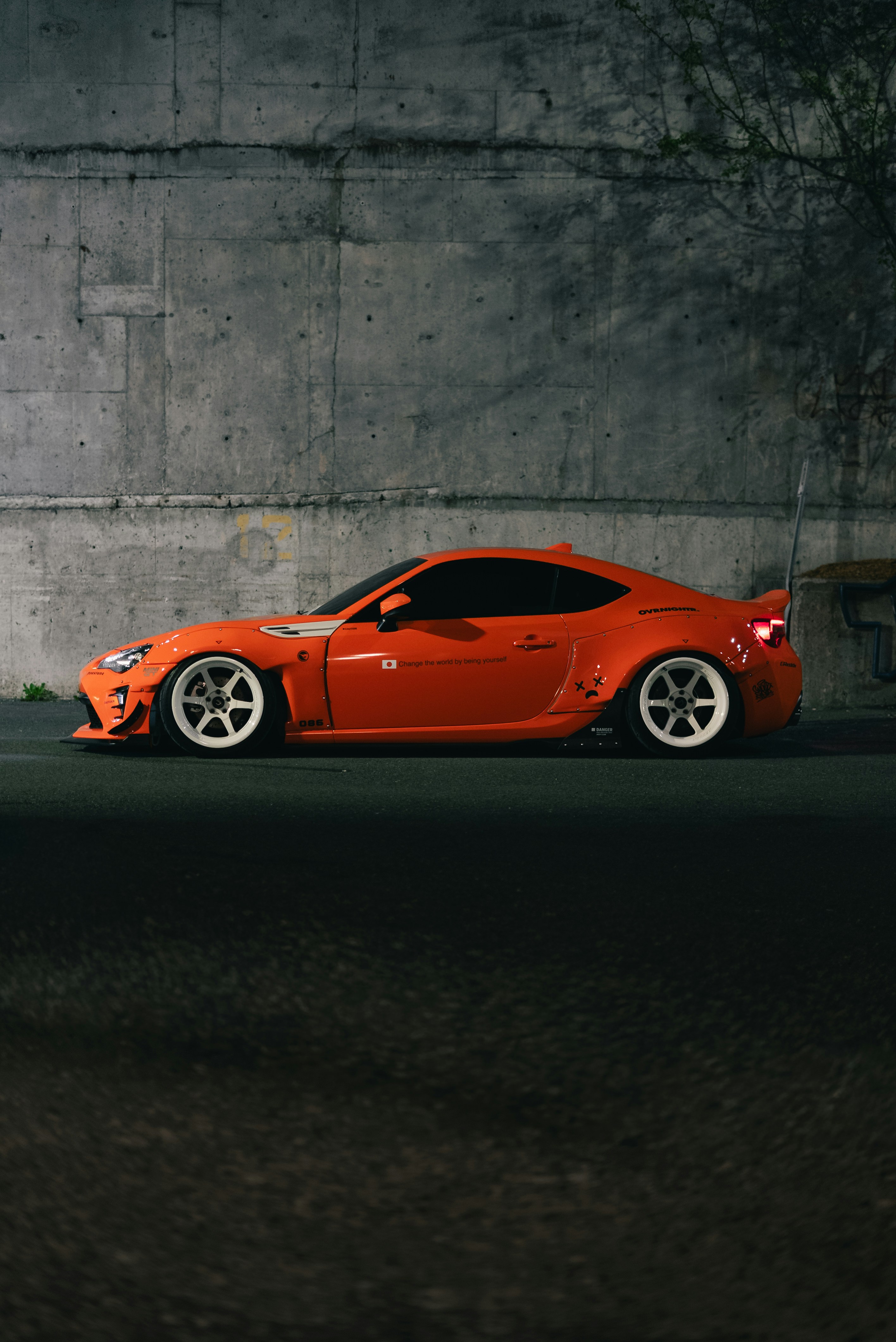 A bright orange sports car with white wheels parked against a concrete wall