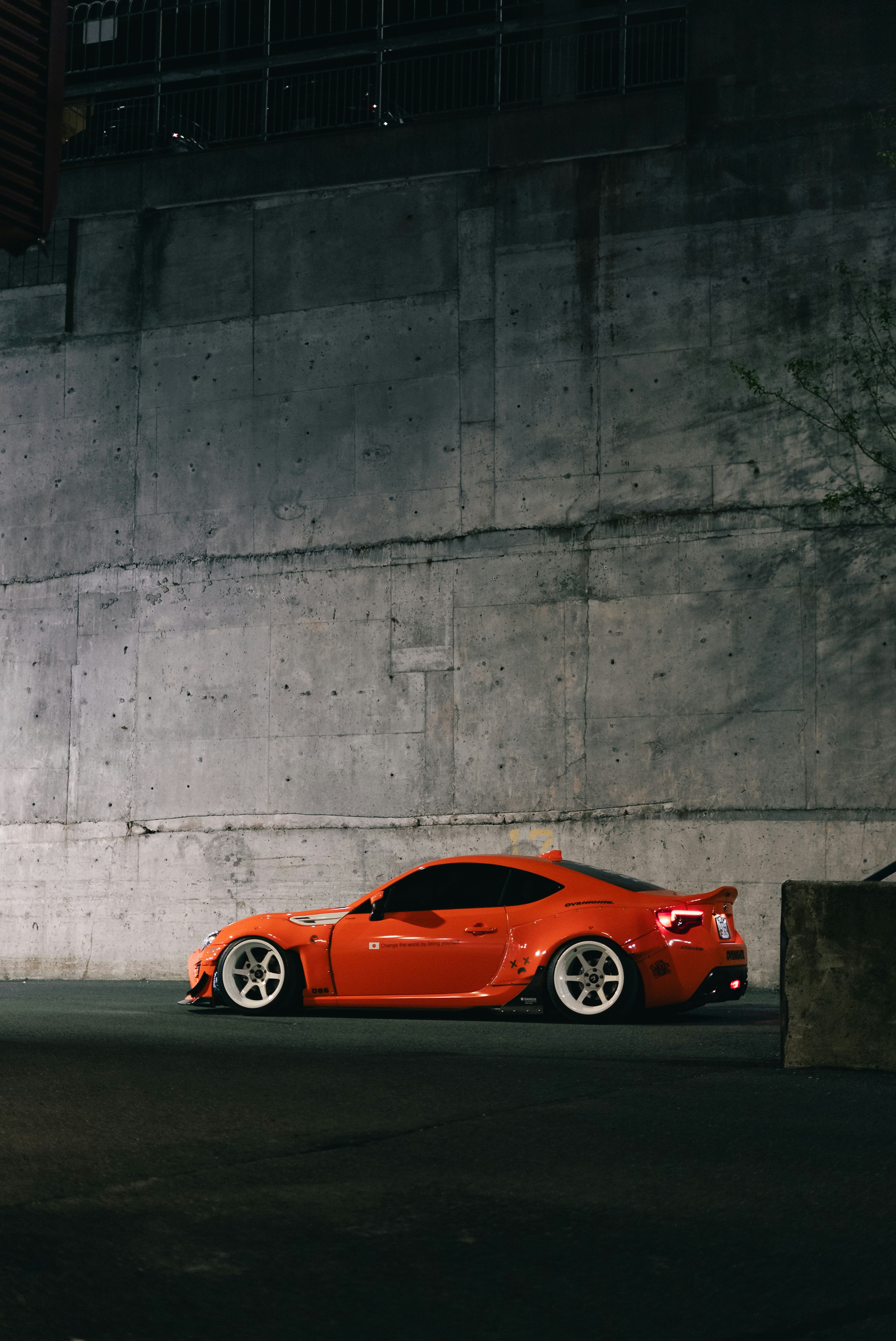 A modified orange sports car with white wheels parked against a concrete wall