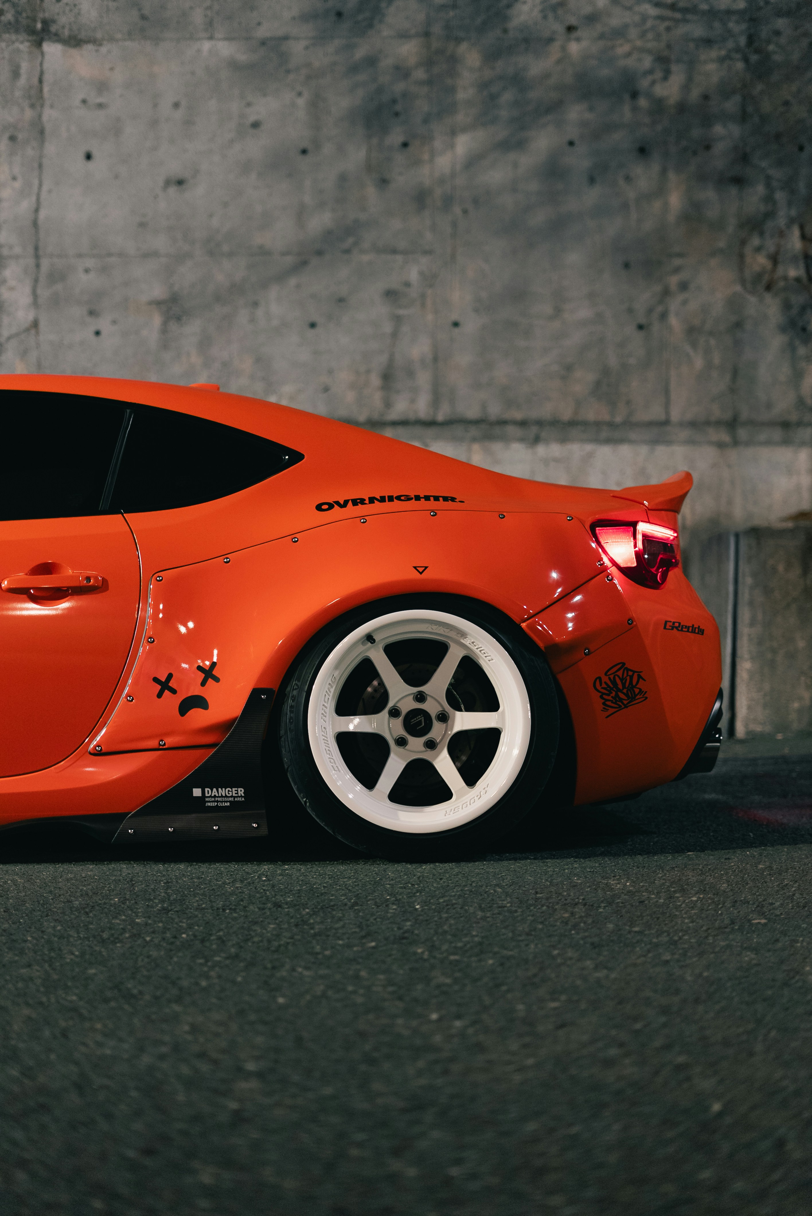 An orange sports car with wide fenders and white wheels against a concrete wall