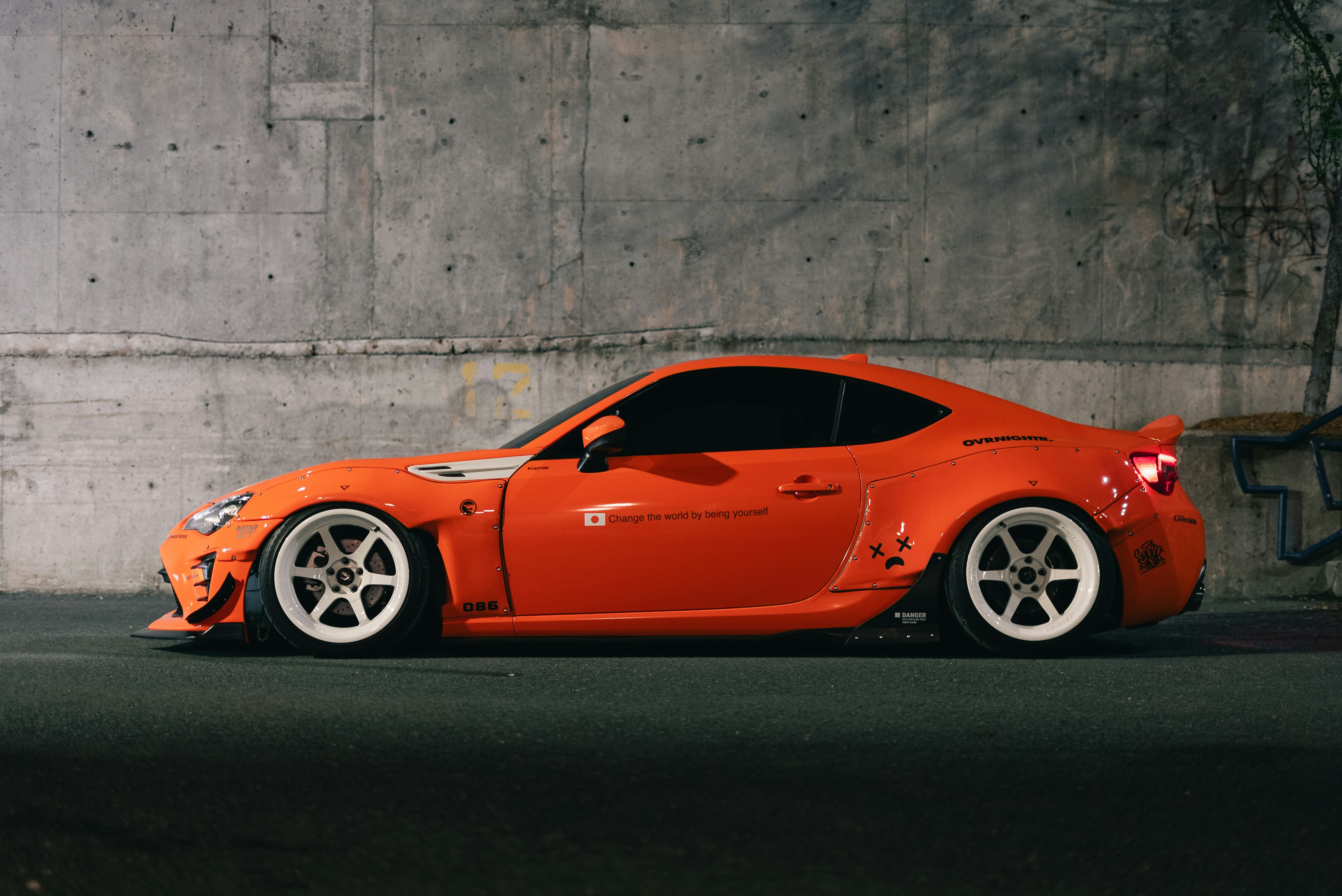 A modified orange sports car with white wheels parked against a concrete wall