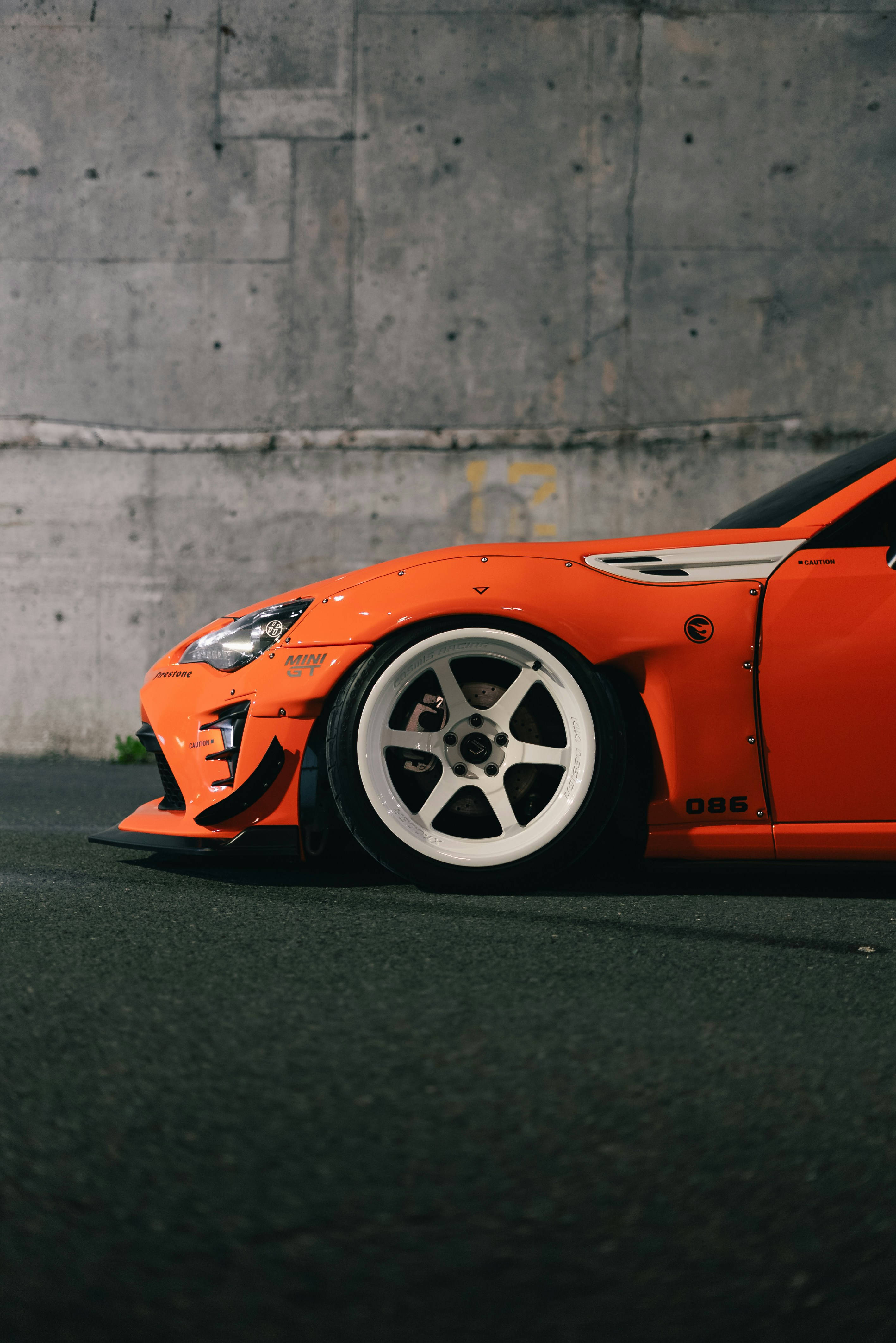A modified orange sports car with white wheels parked against a concrete wall