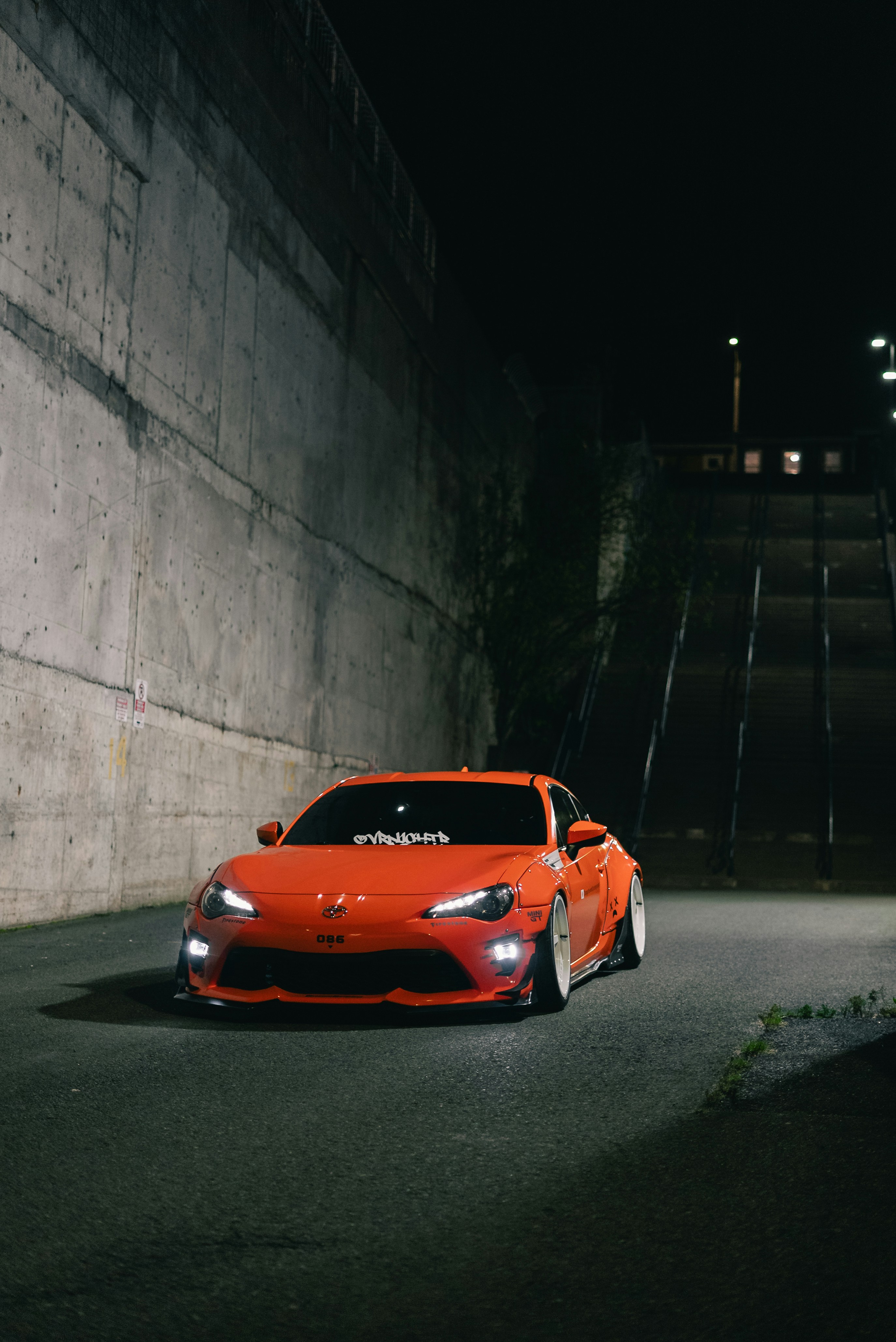 An orange sports car parked on a dark street beside a concrete wall