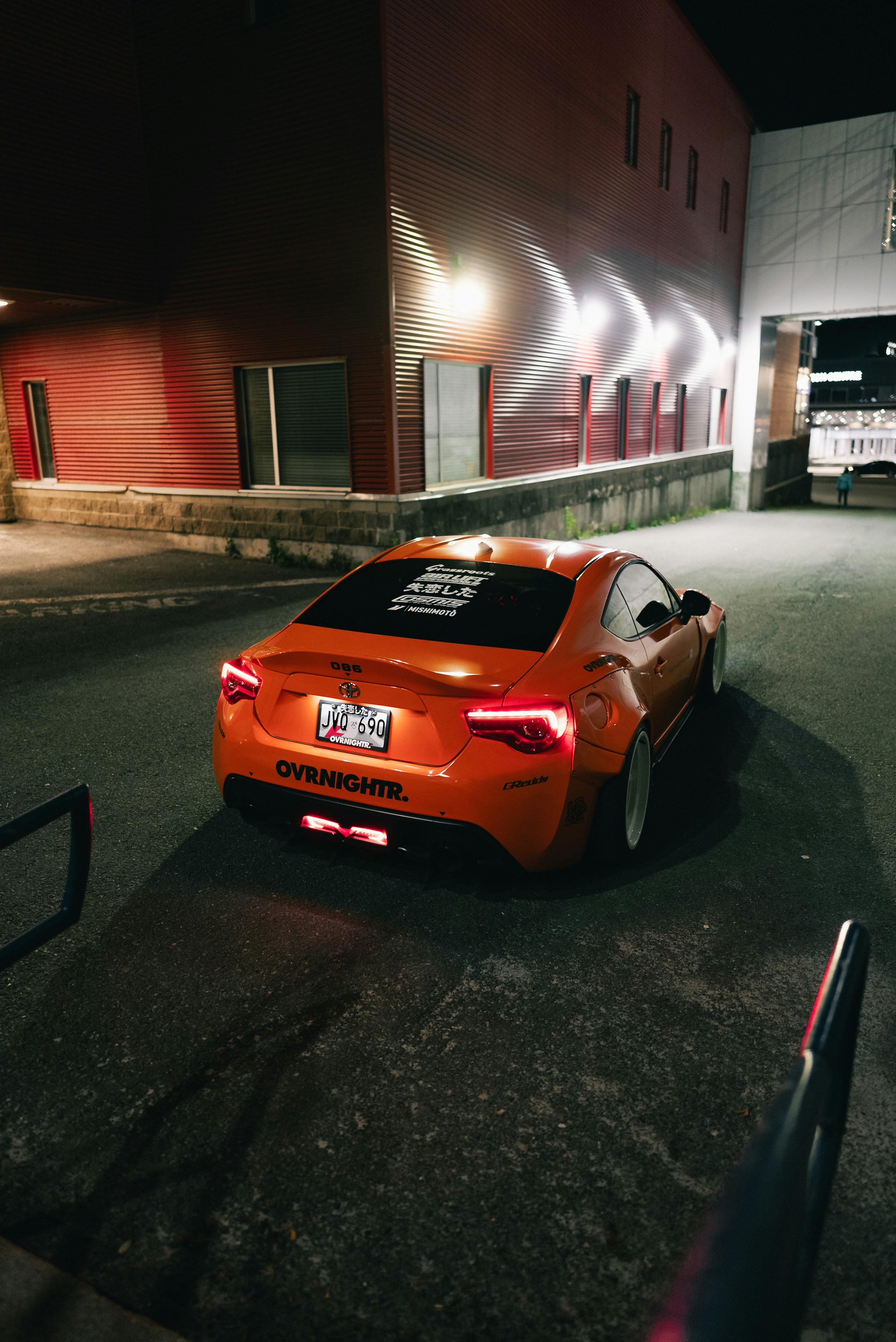 An orange sports car parked at night beside a red corrugated metal building