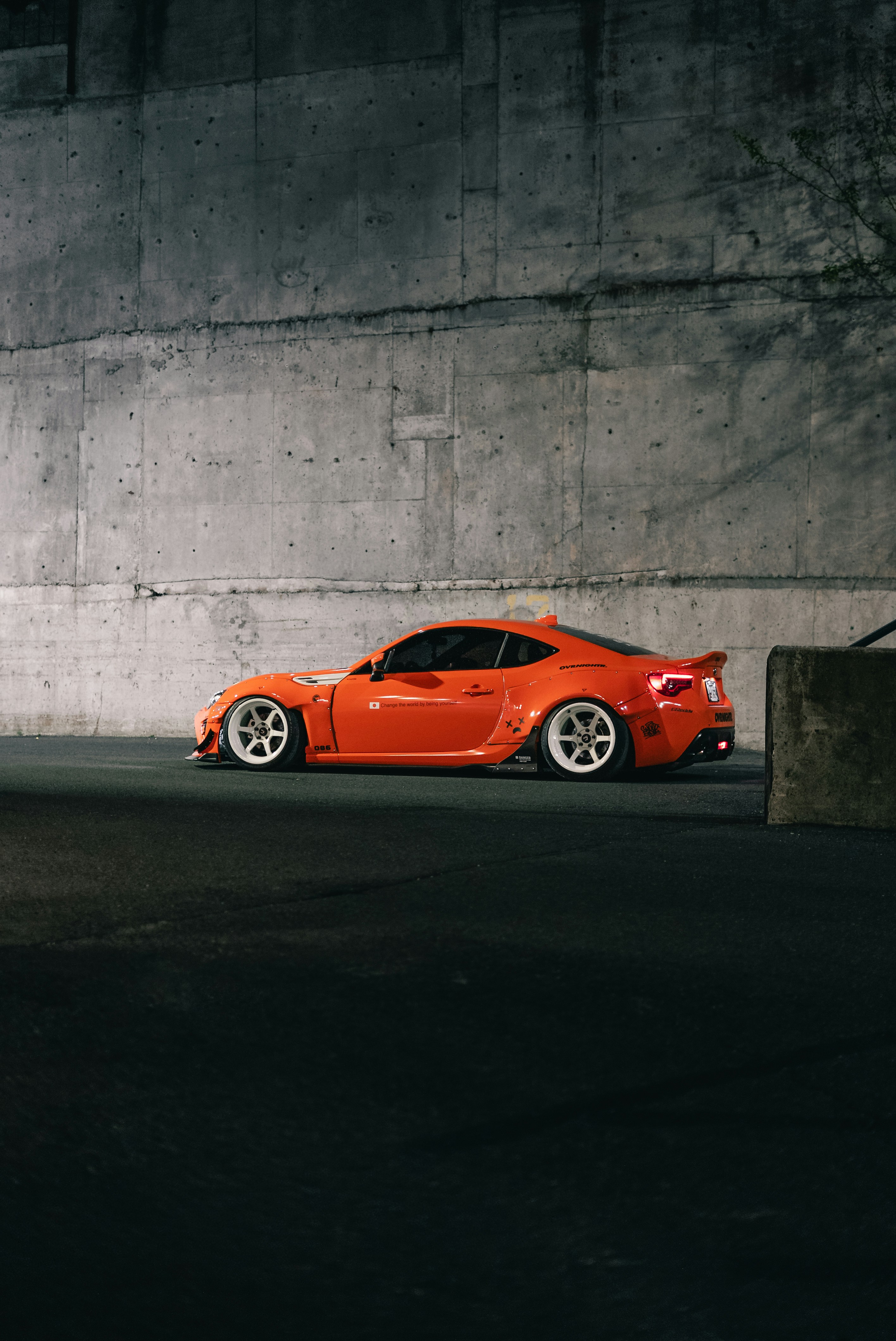 An orange sports car with white wheels parked against a tall concrete wall