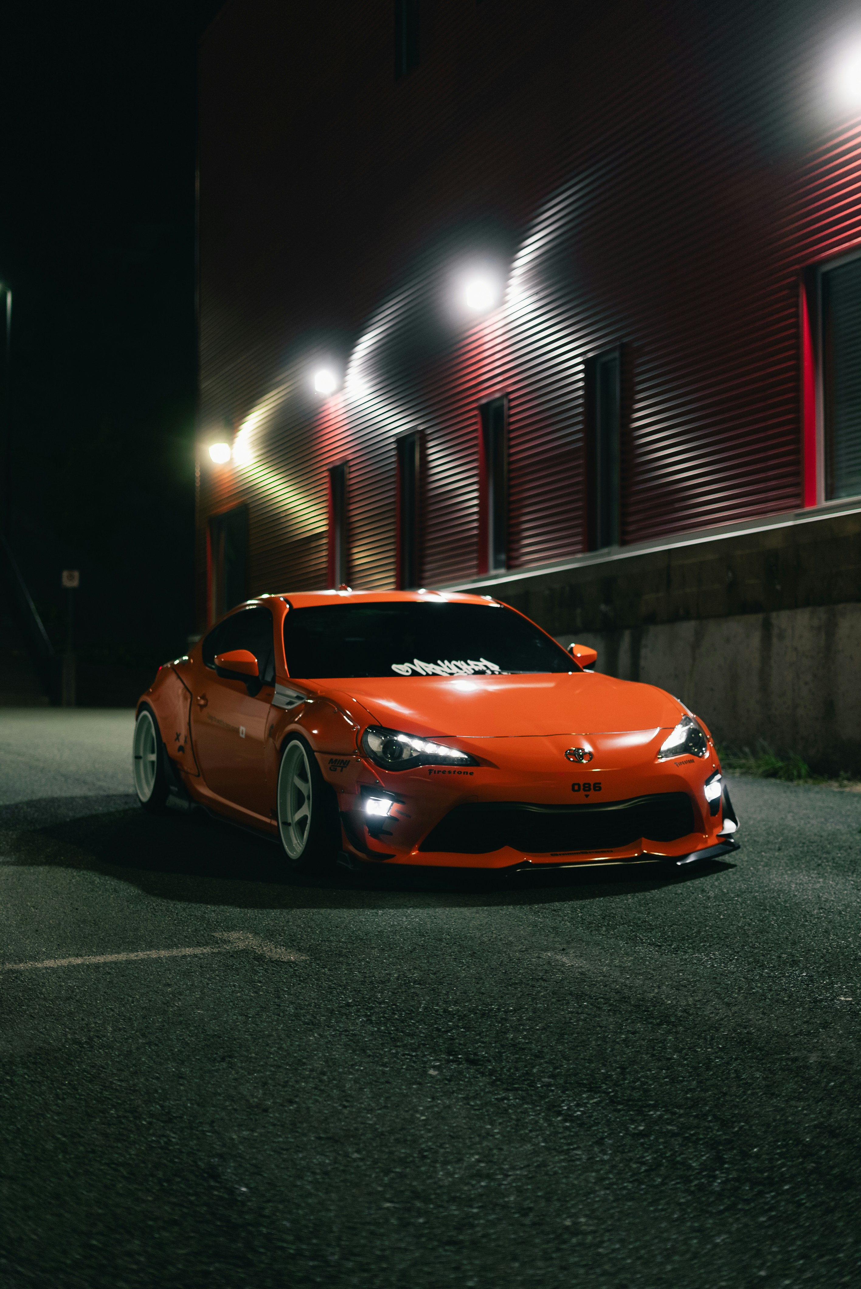 An orange sports car with white wheels parked beside a red industrial building