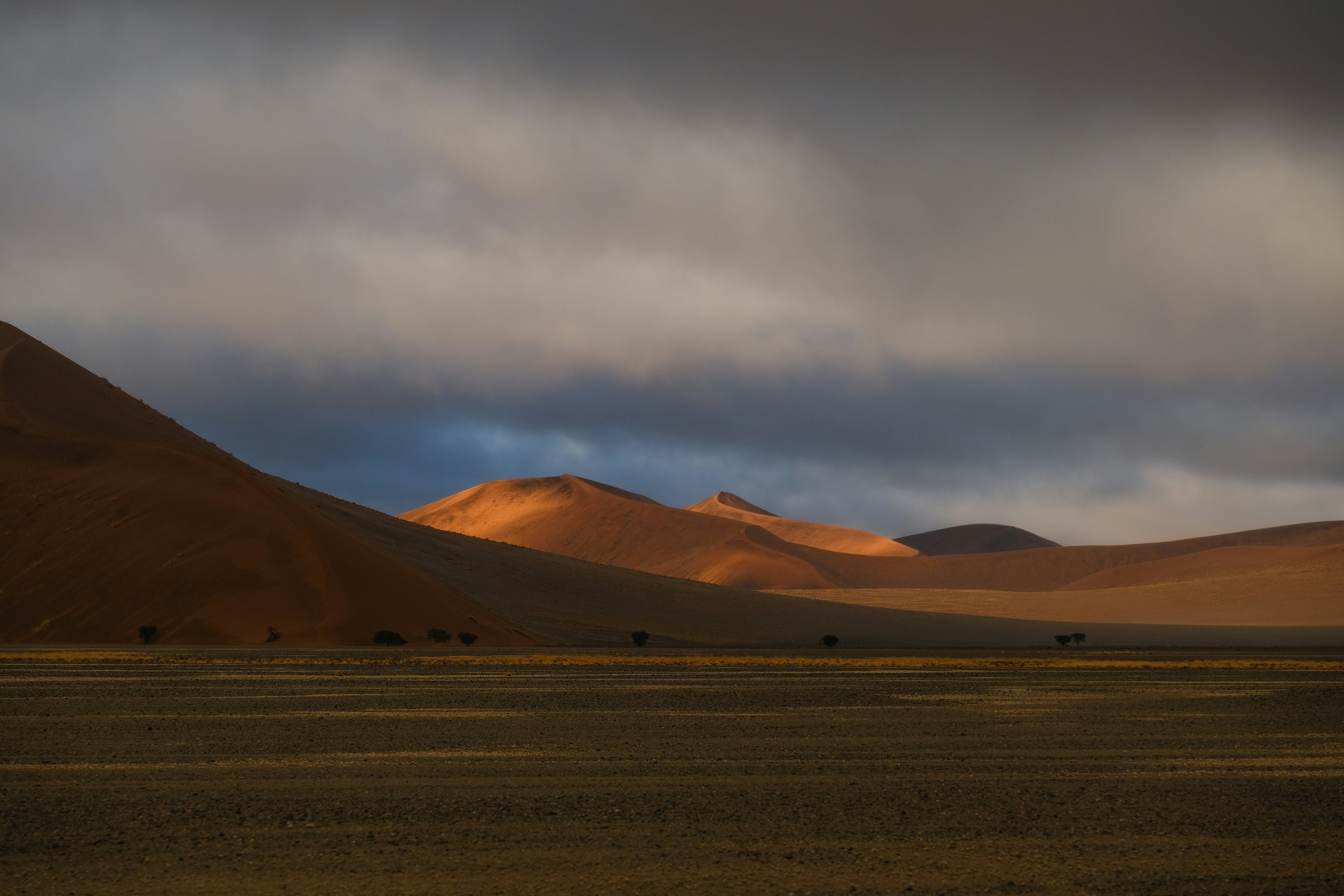 Orange sand dunes under a dark cloudy sky in a vast desert landscape