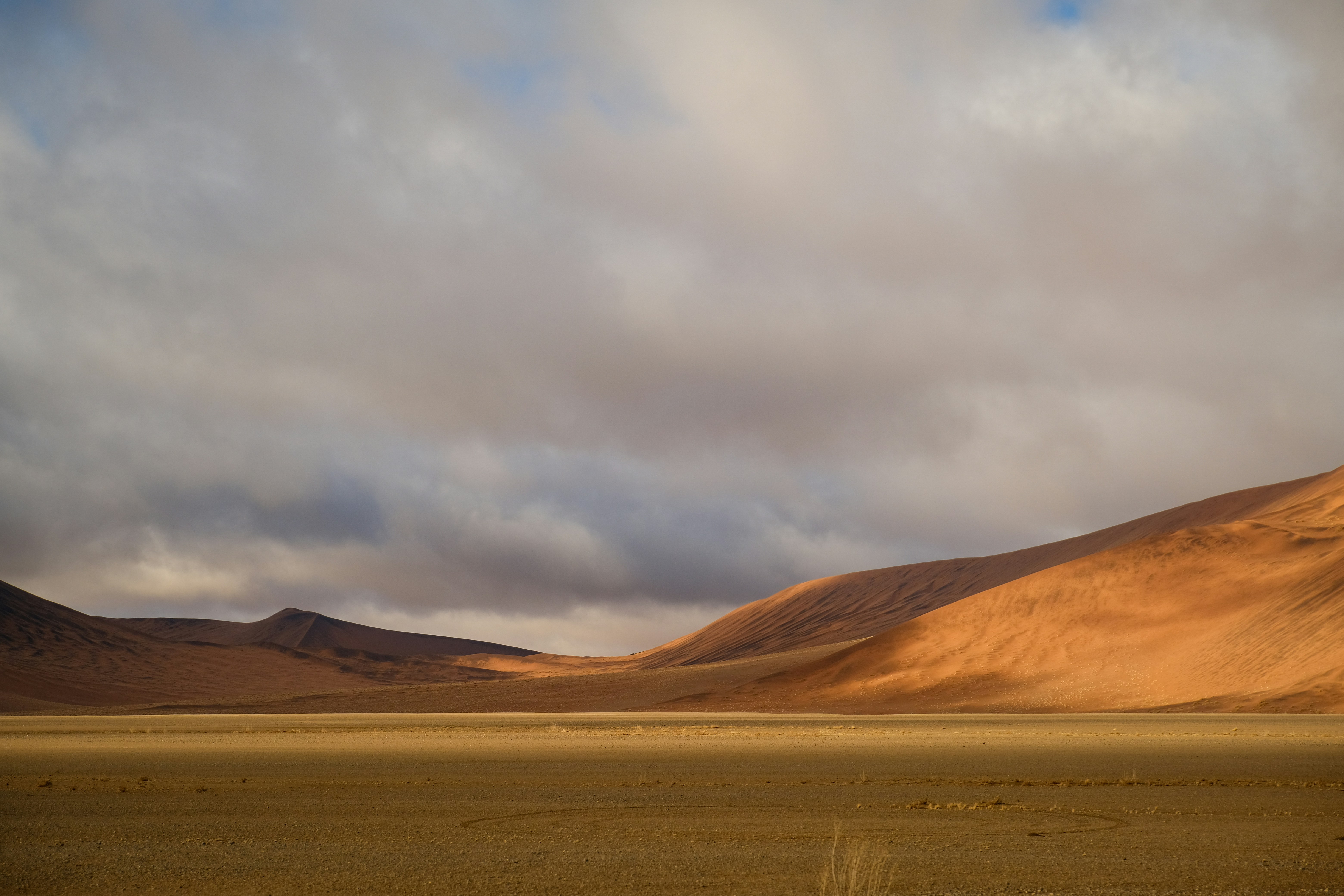 Orange sand dunes rising above a flat desert floor under a cloudy sky