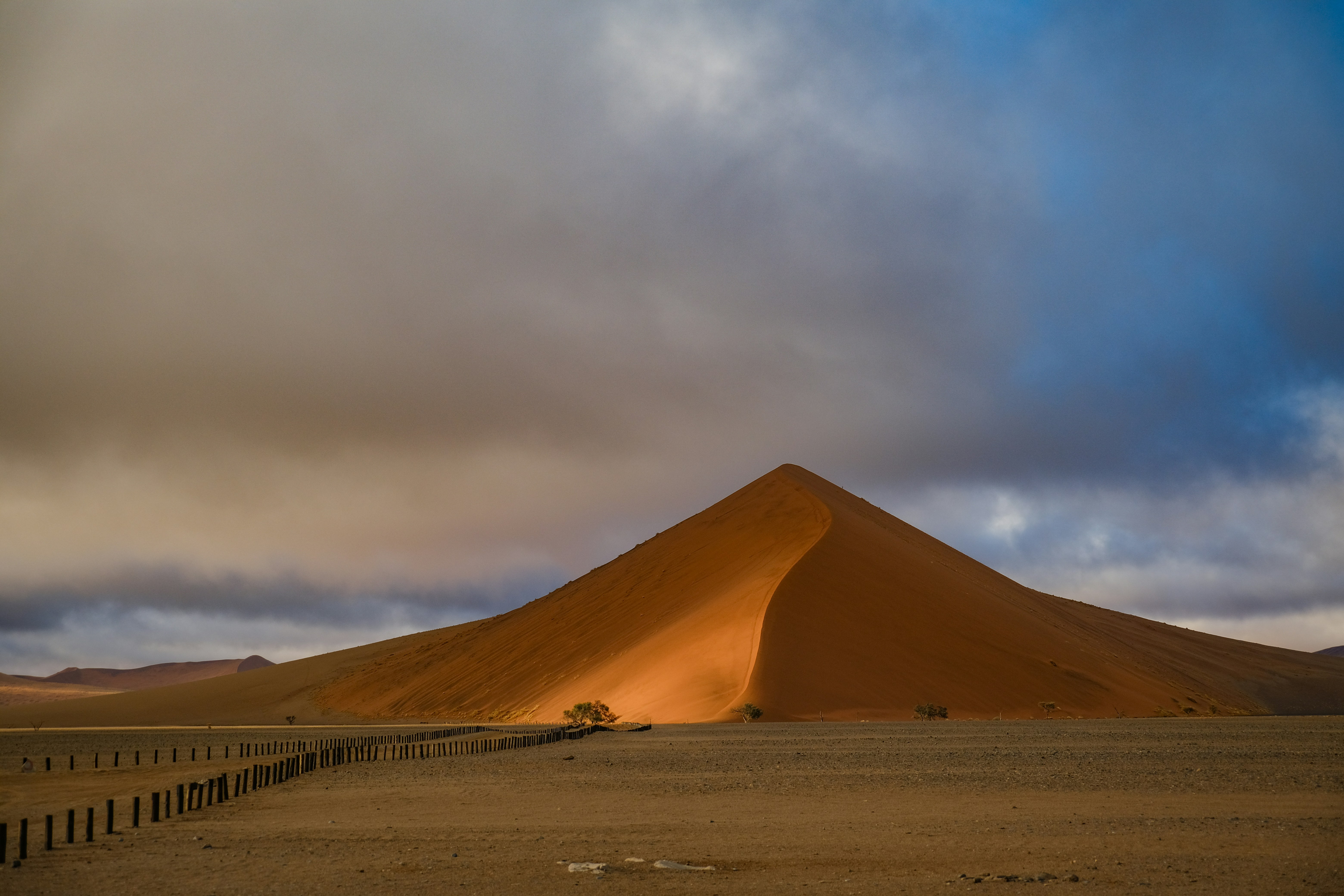A large orange sand dune under a cloudy sky with a wooden fence