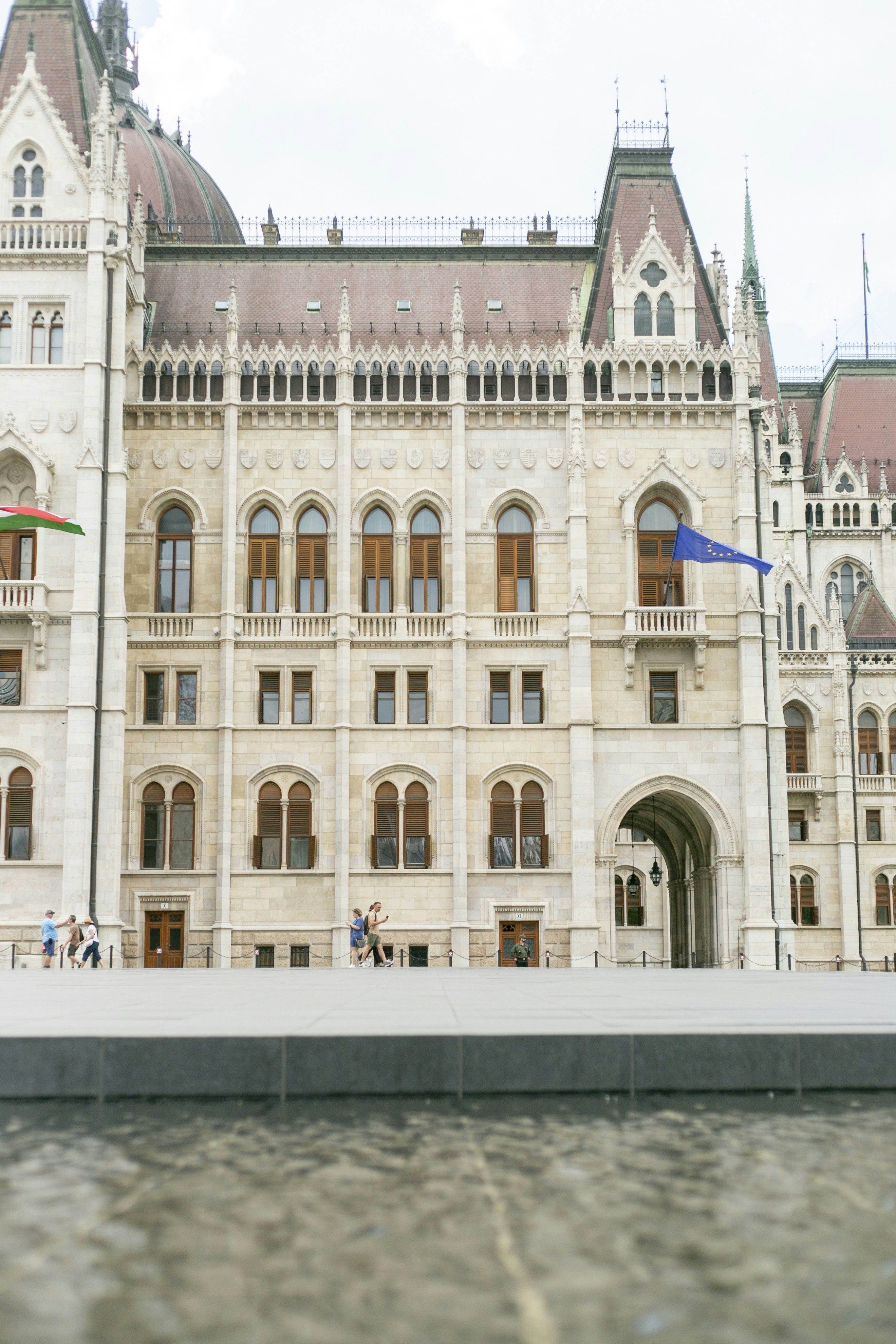 People walking past the ornate stone facade of the Hungarian Parliament Building in Budapest