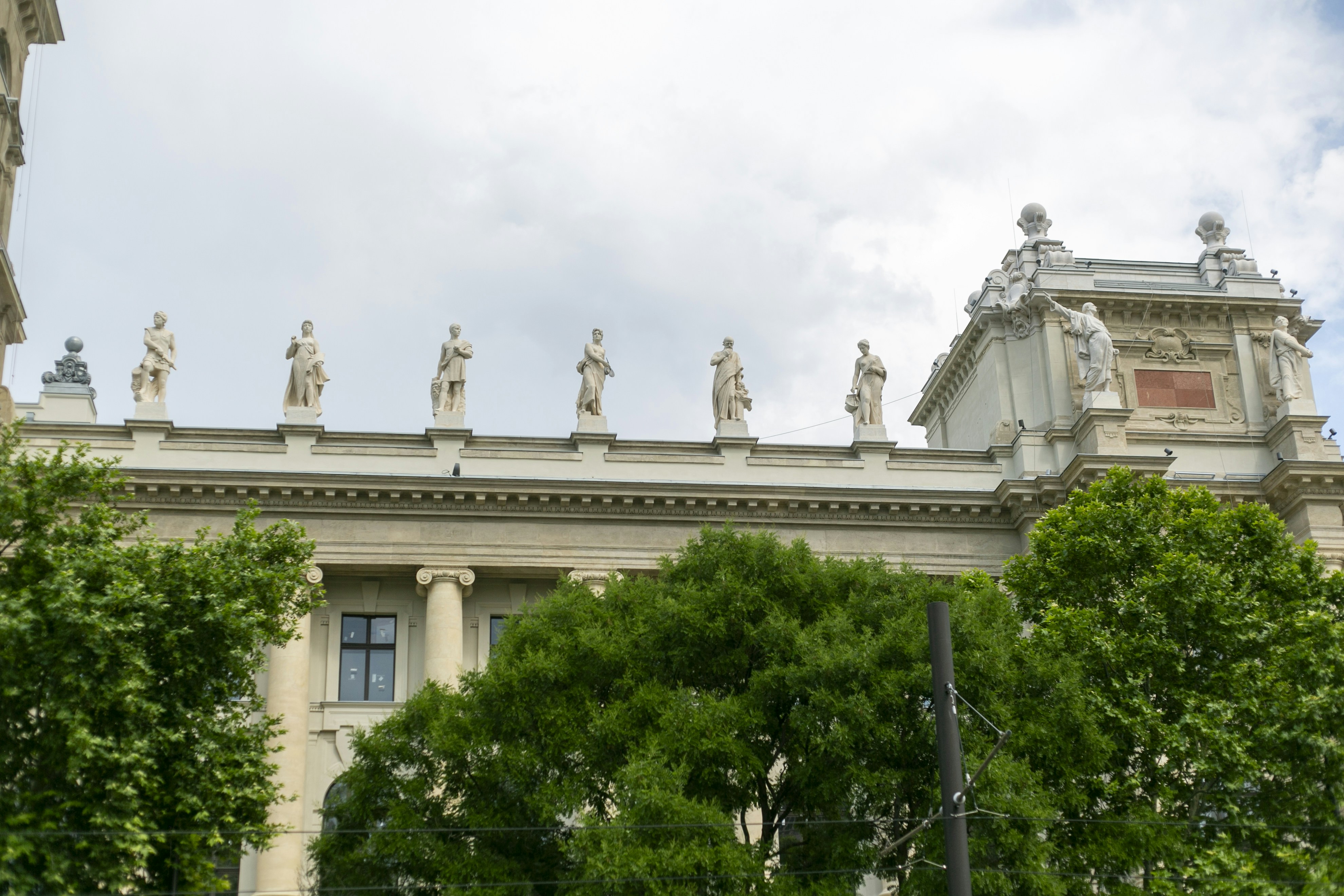 Stone statues lining the roof of a neoclassical building behind green trees