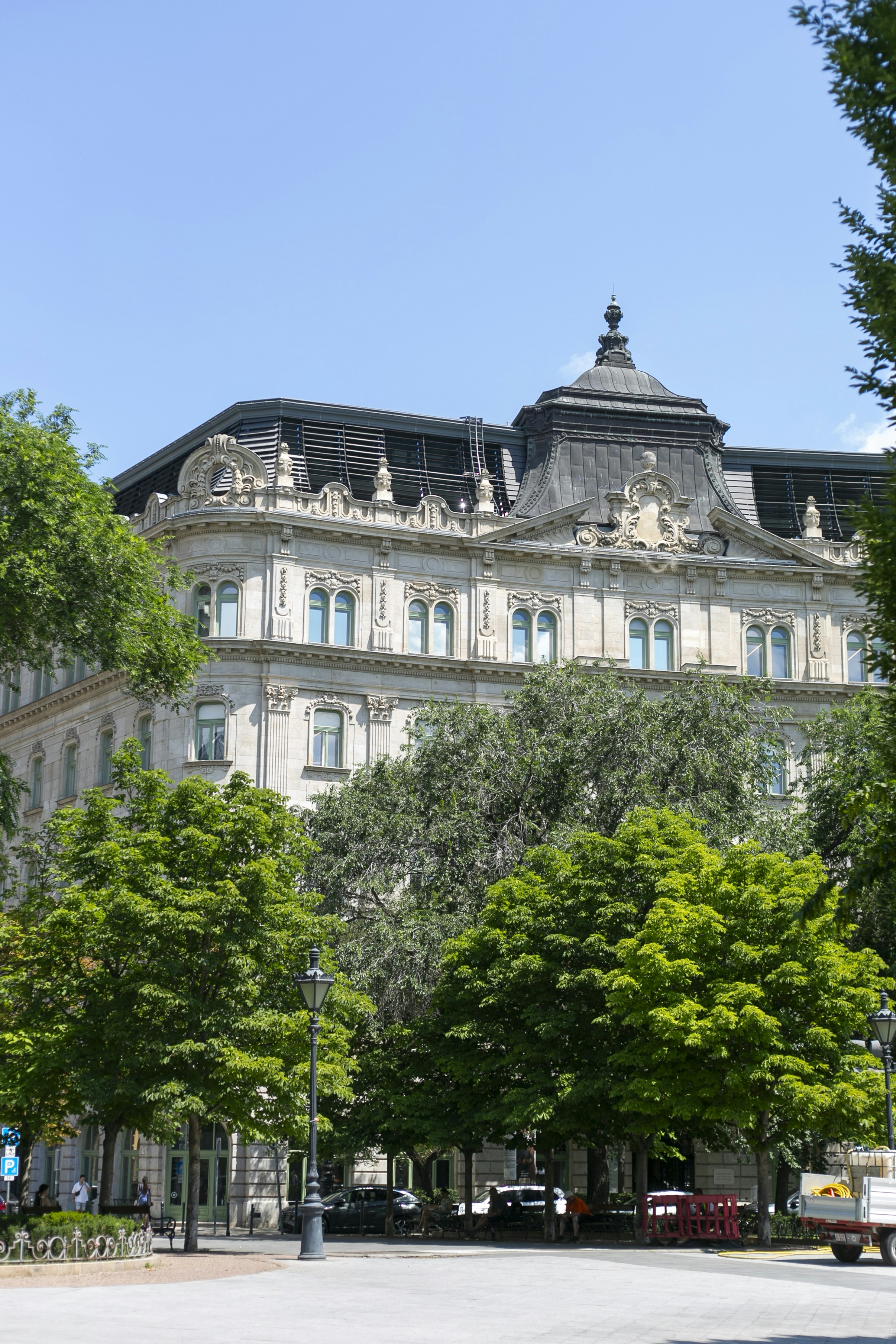 An ornate stone building with a dark mansard roof behind lush green trees