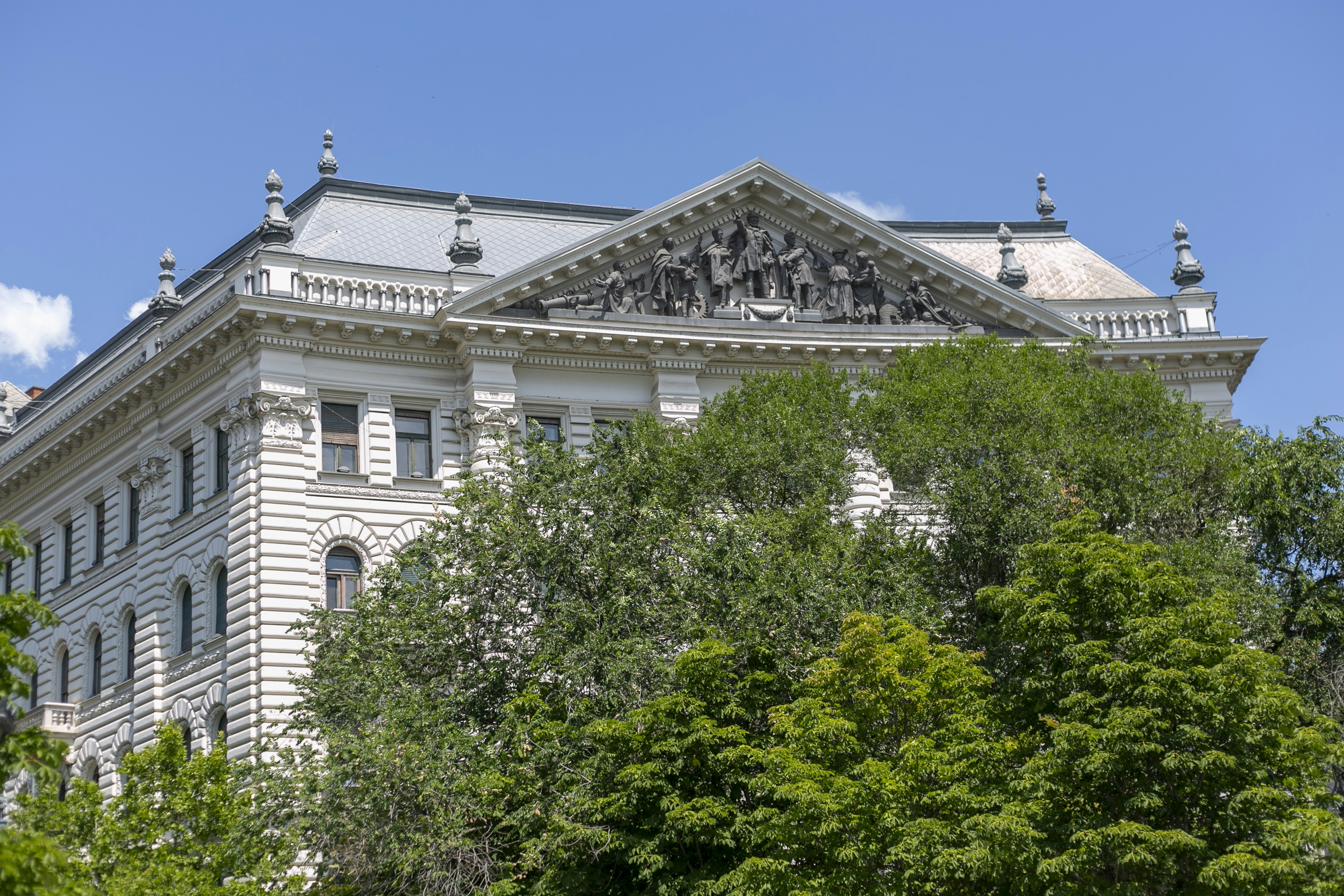 A neoclassical building with a sculpted pediment behind lush green trees under blue sky