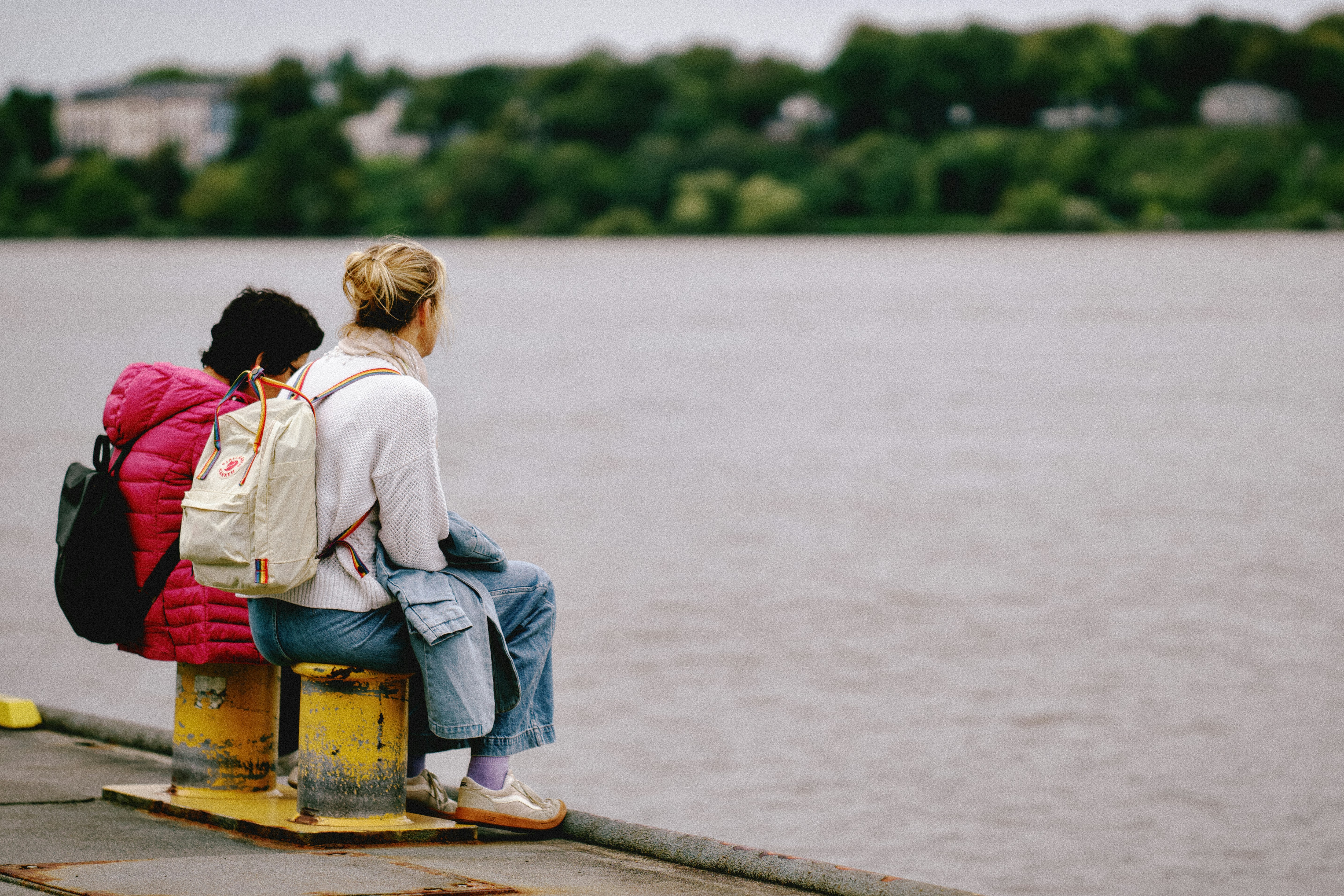 Two people with backpacks sitting on yellow bollards by a wide river