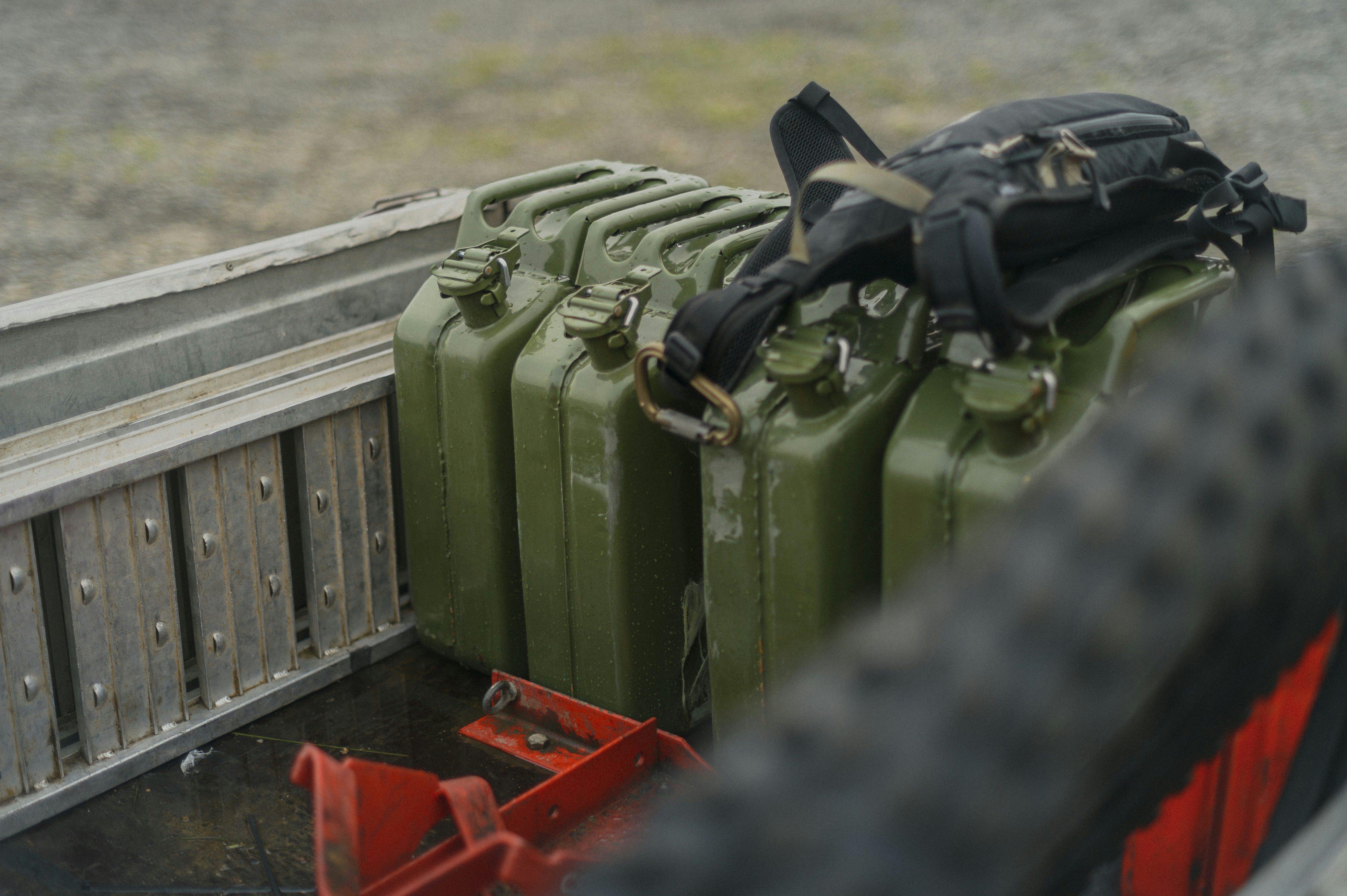 Row of green metal fuel cans and a black backpack in a pickup bed, with a red tie-down bracket in the foreground