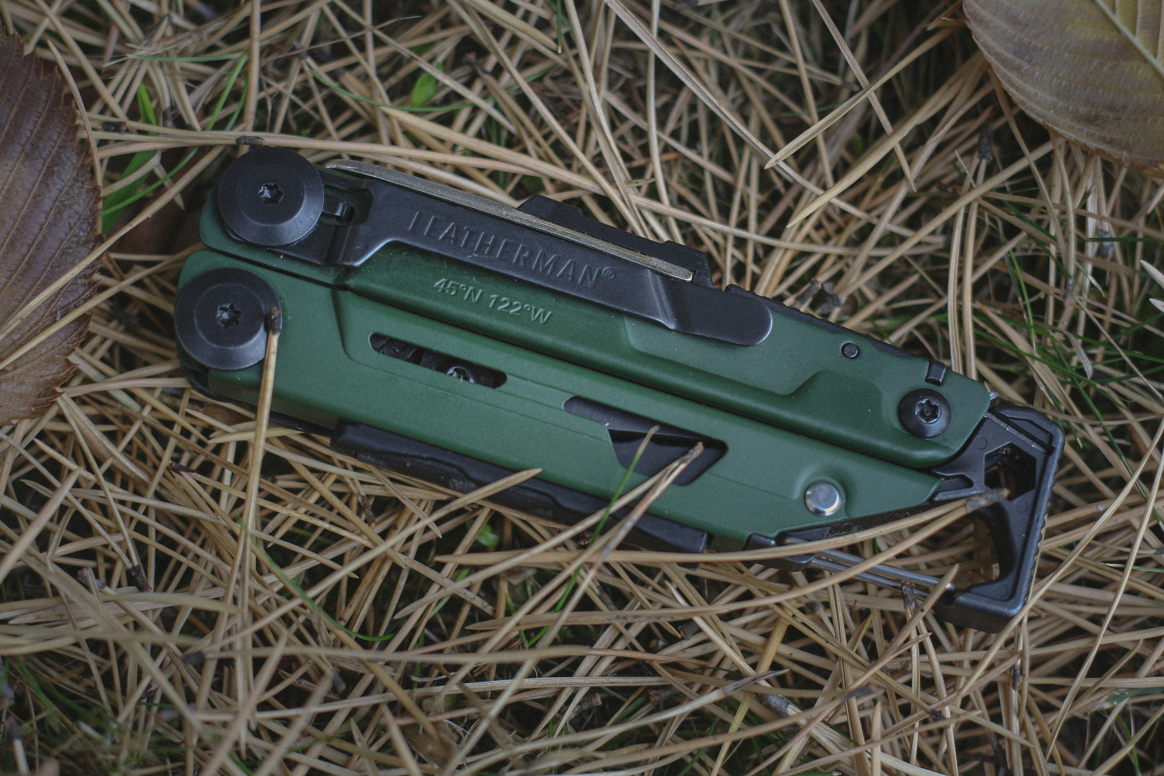 A green multi-tool resting on a bed of dry pine needles