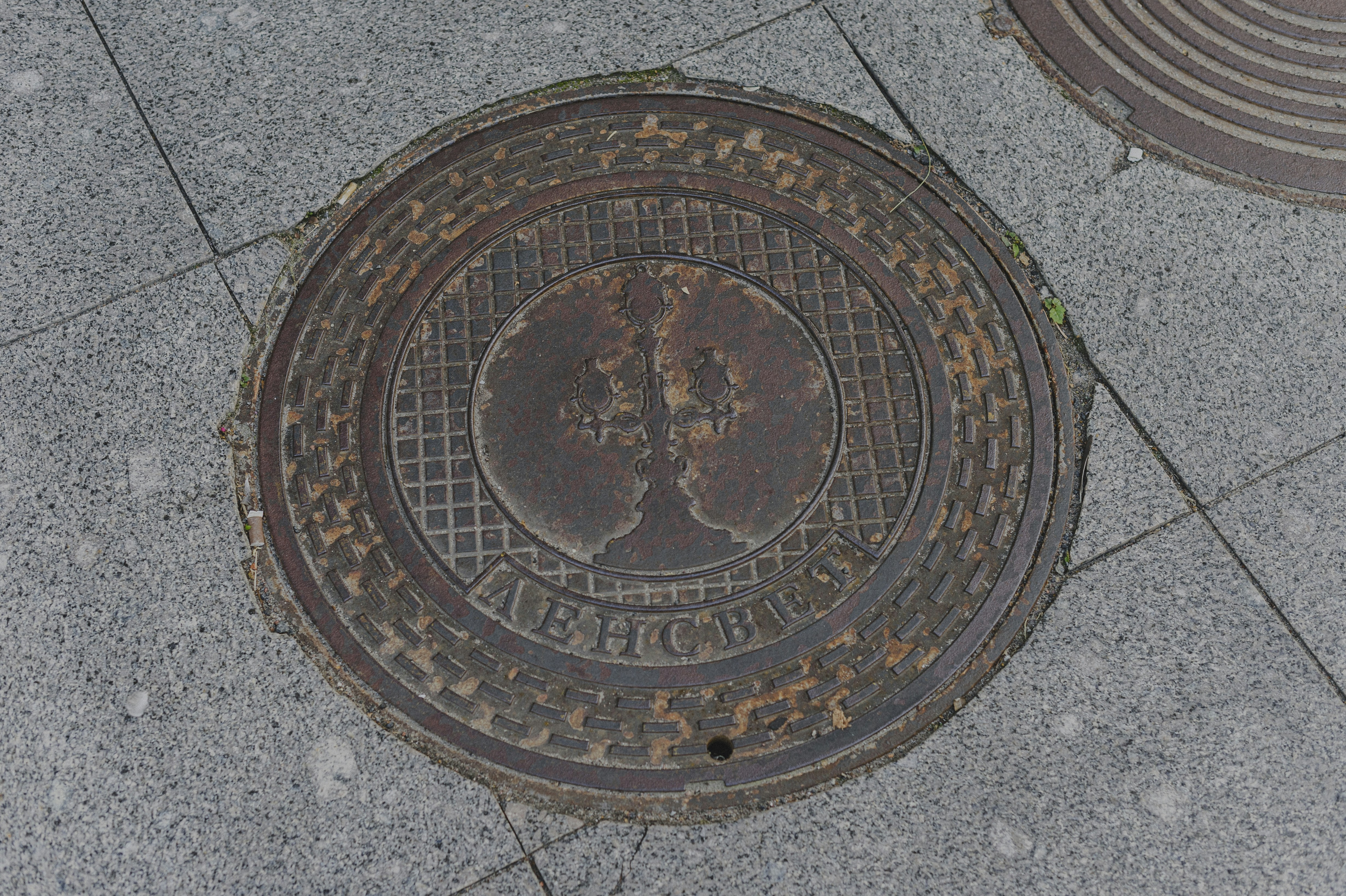 A circular metal manhole cover with a candelabra emblem on a grey stone sidewalk
