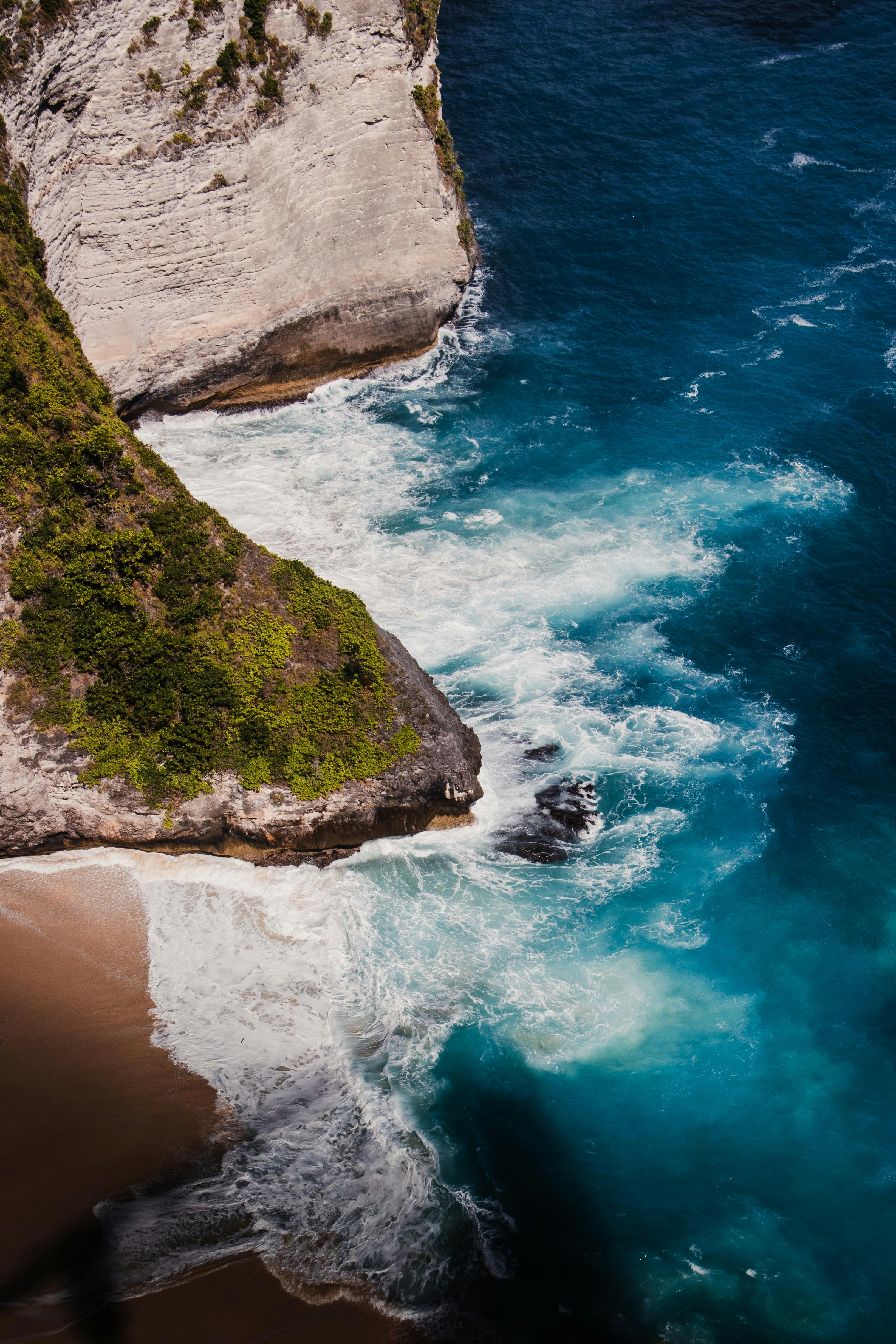 Waves crashing against a steep cliff with green vegetation above a sandy beach