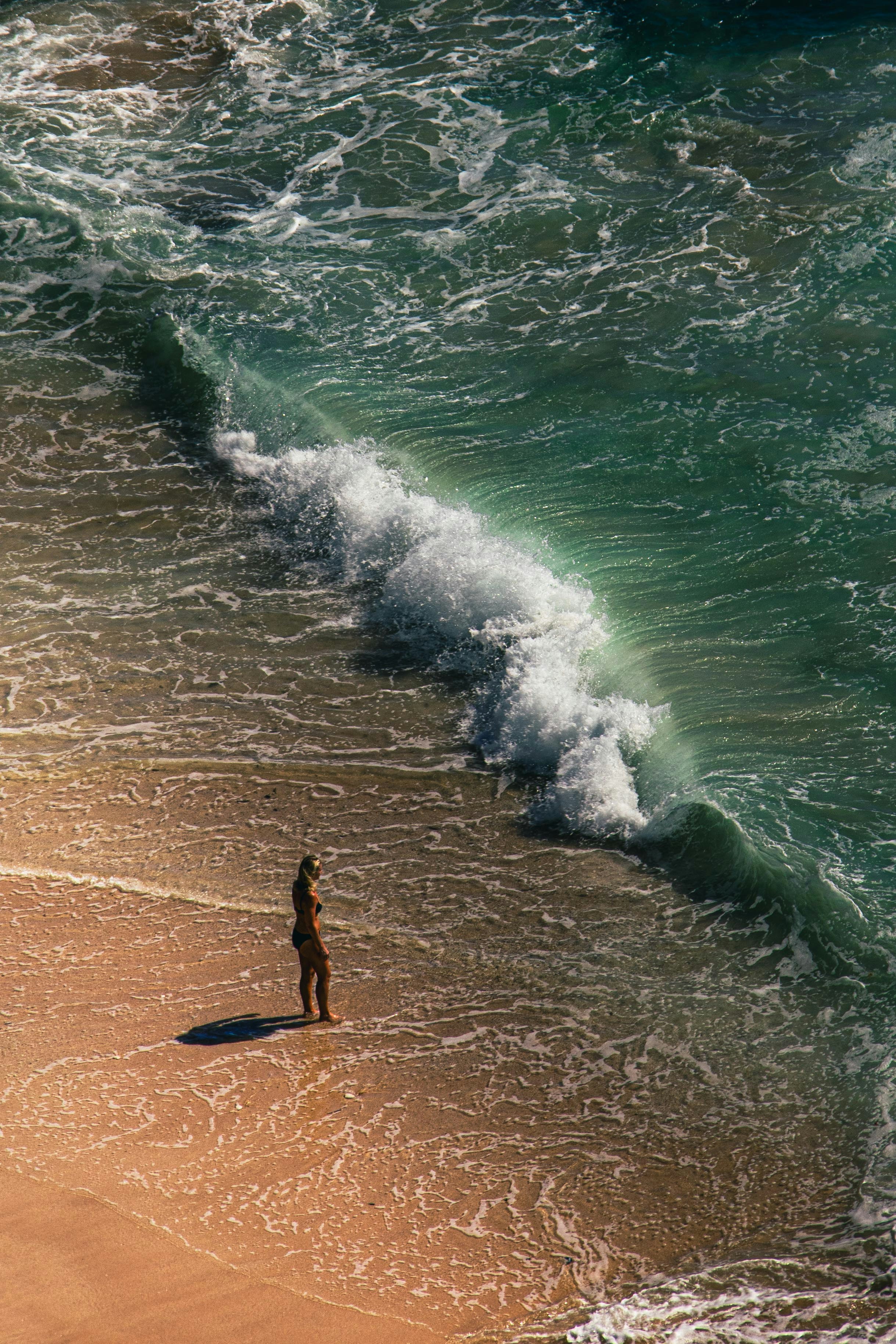 A woman in a bikini standing on a sandy beach near a breaking wave