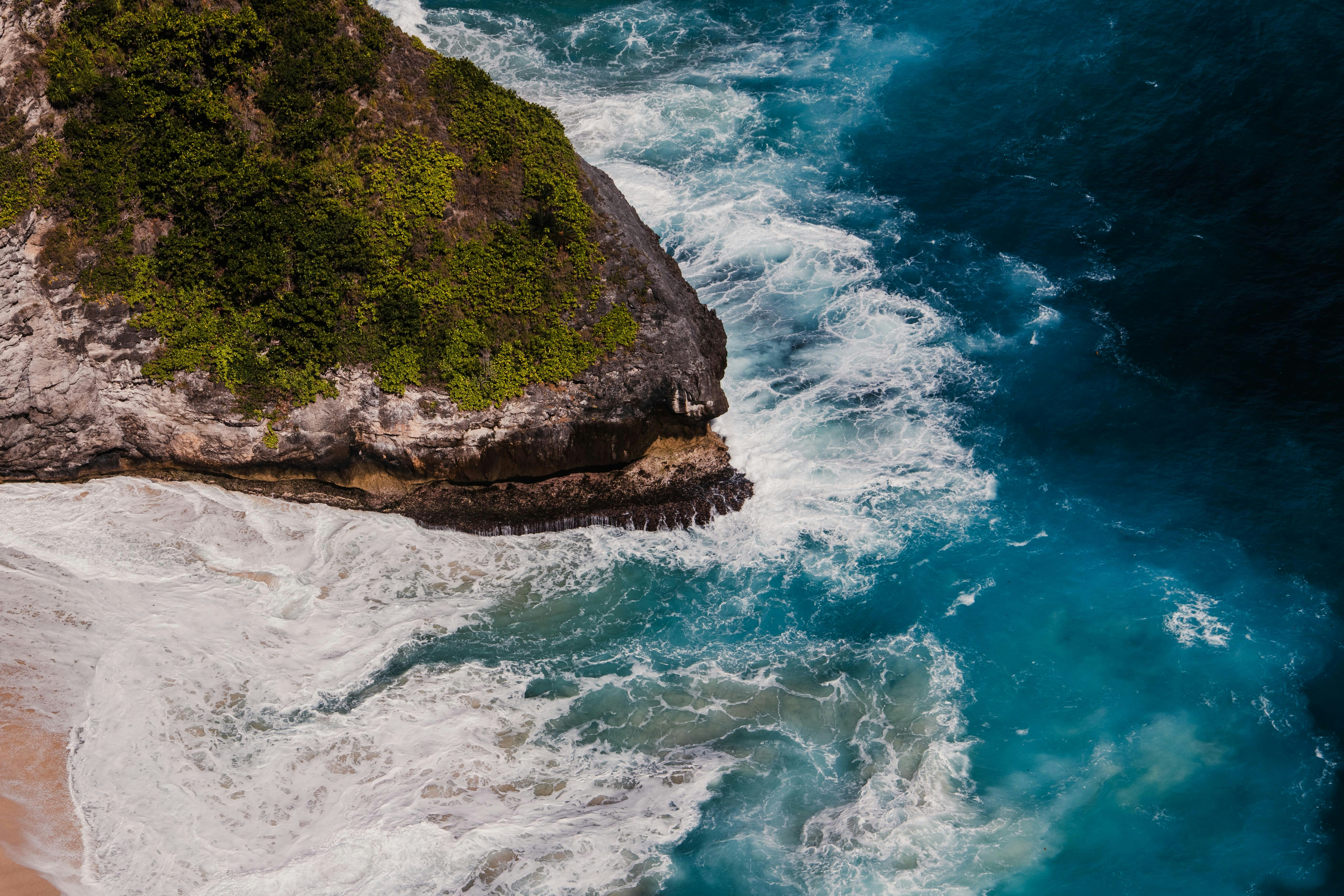 Aerial view of turquoise waves crashing against a rocky cliff covered in green vegetation
