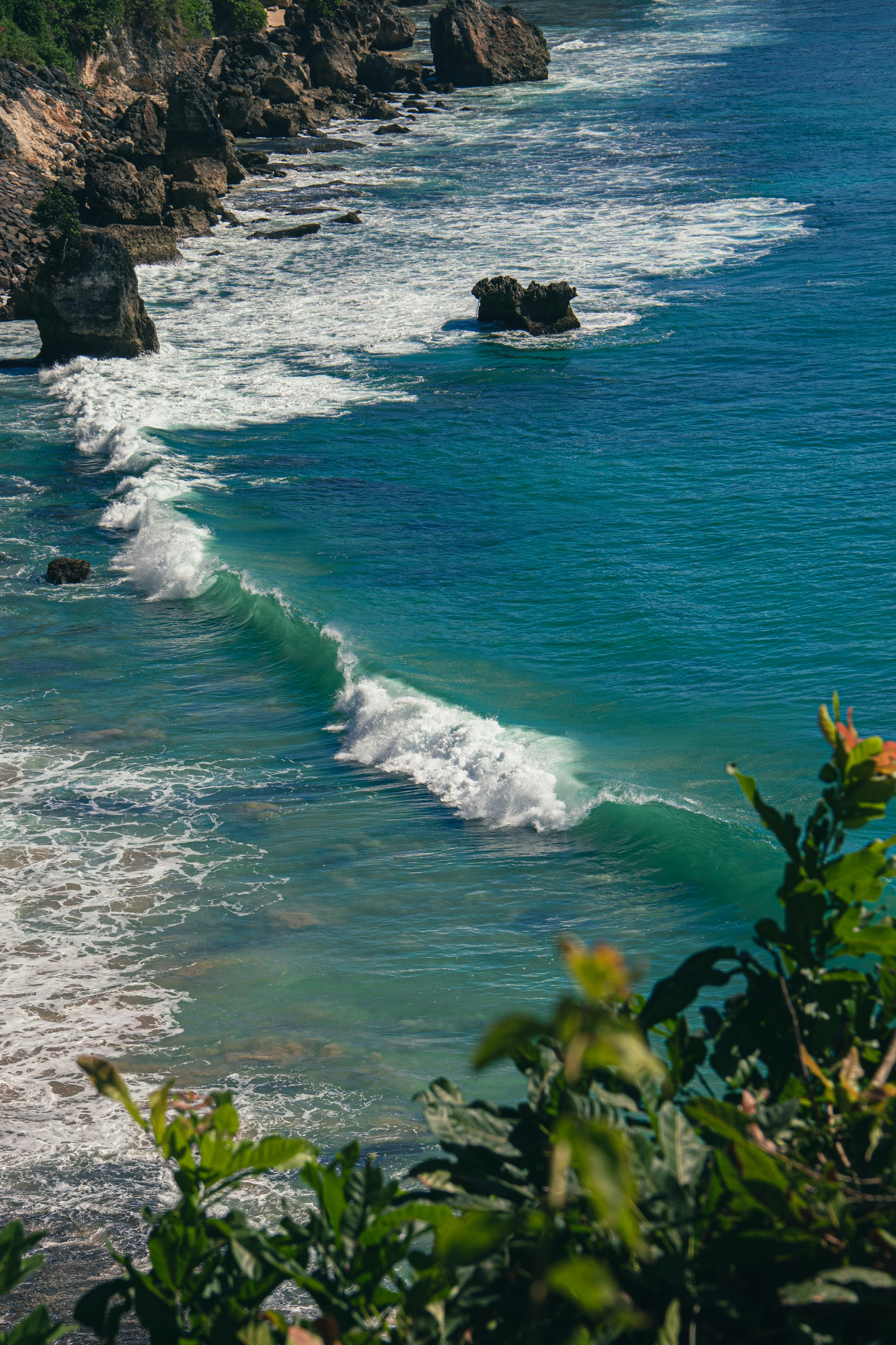 Turquoise ocean waves breaking against a rocky coastline with green foliage in foreground