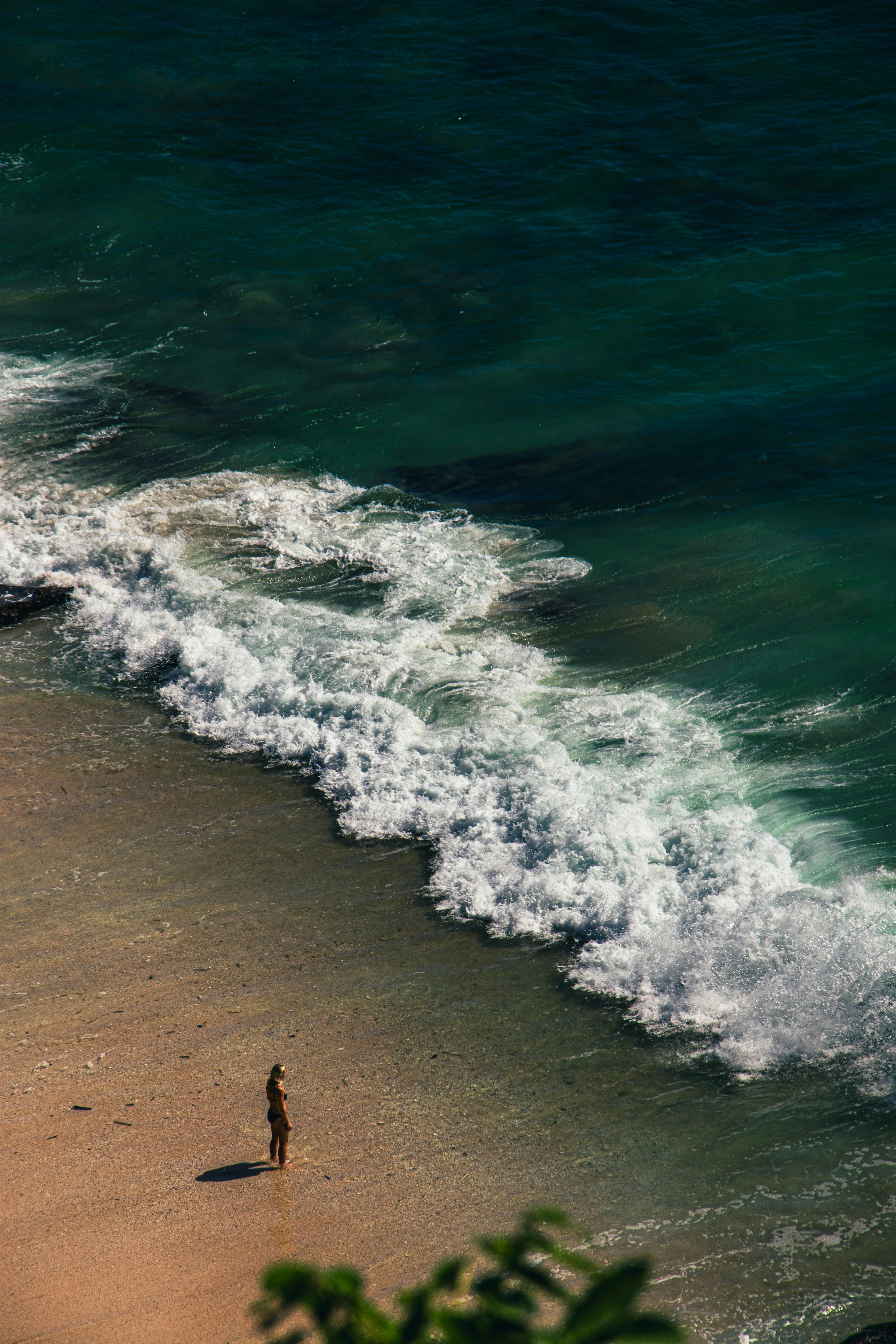 A person in a bikini standing on a sandy beach near crashing waves