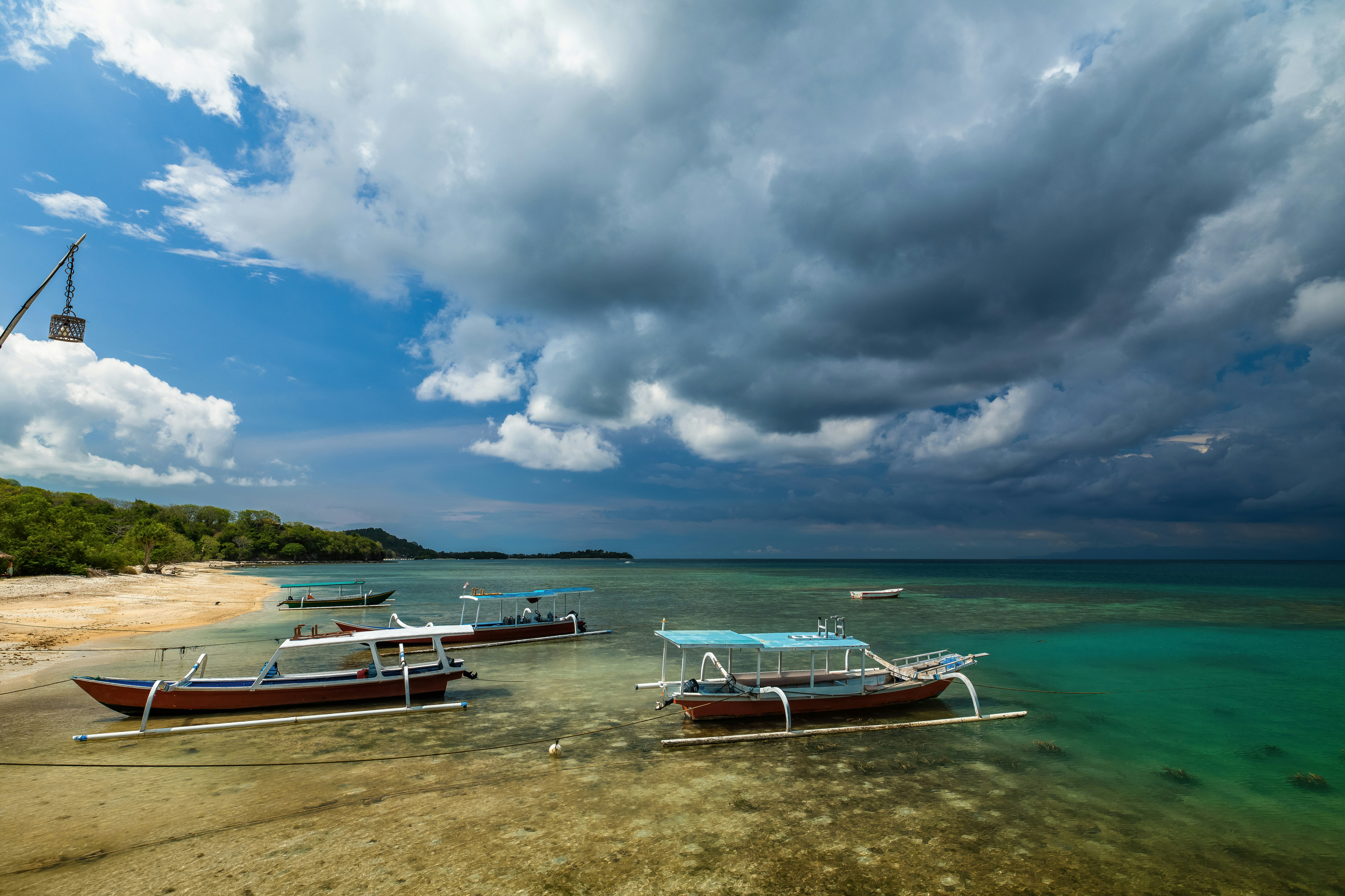 Outrigger boats anchored in shallow turquoise water near a sandy beach under stormy clouds