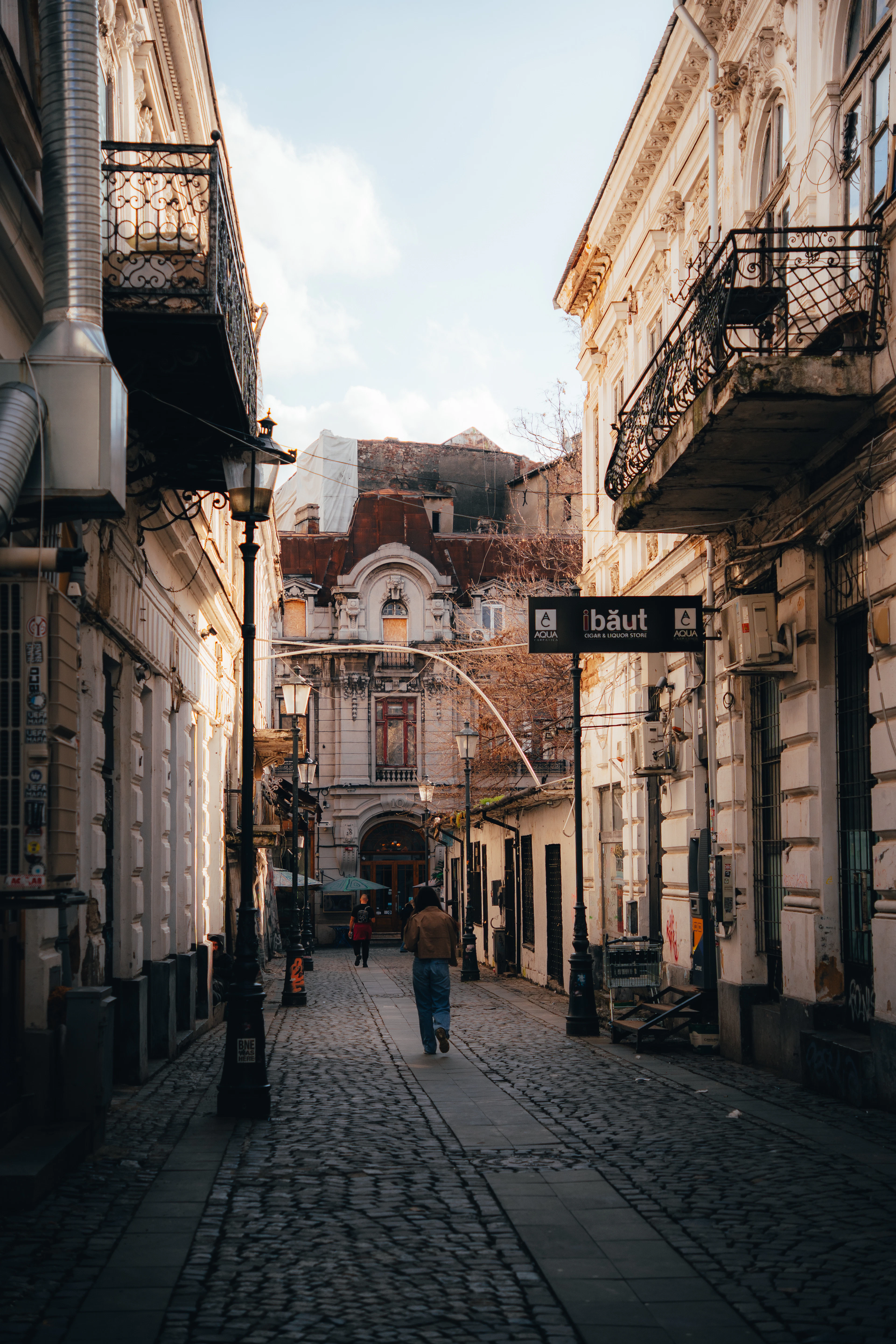 Warm evening light fills a cobblestone Old Town alley lined with wrought-iron balconies as a lone walker heads down it.